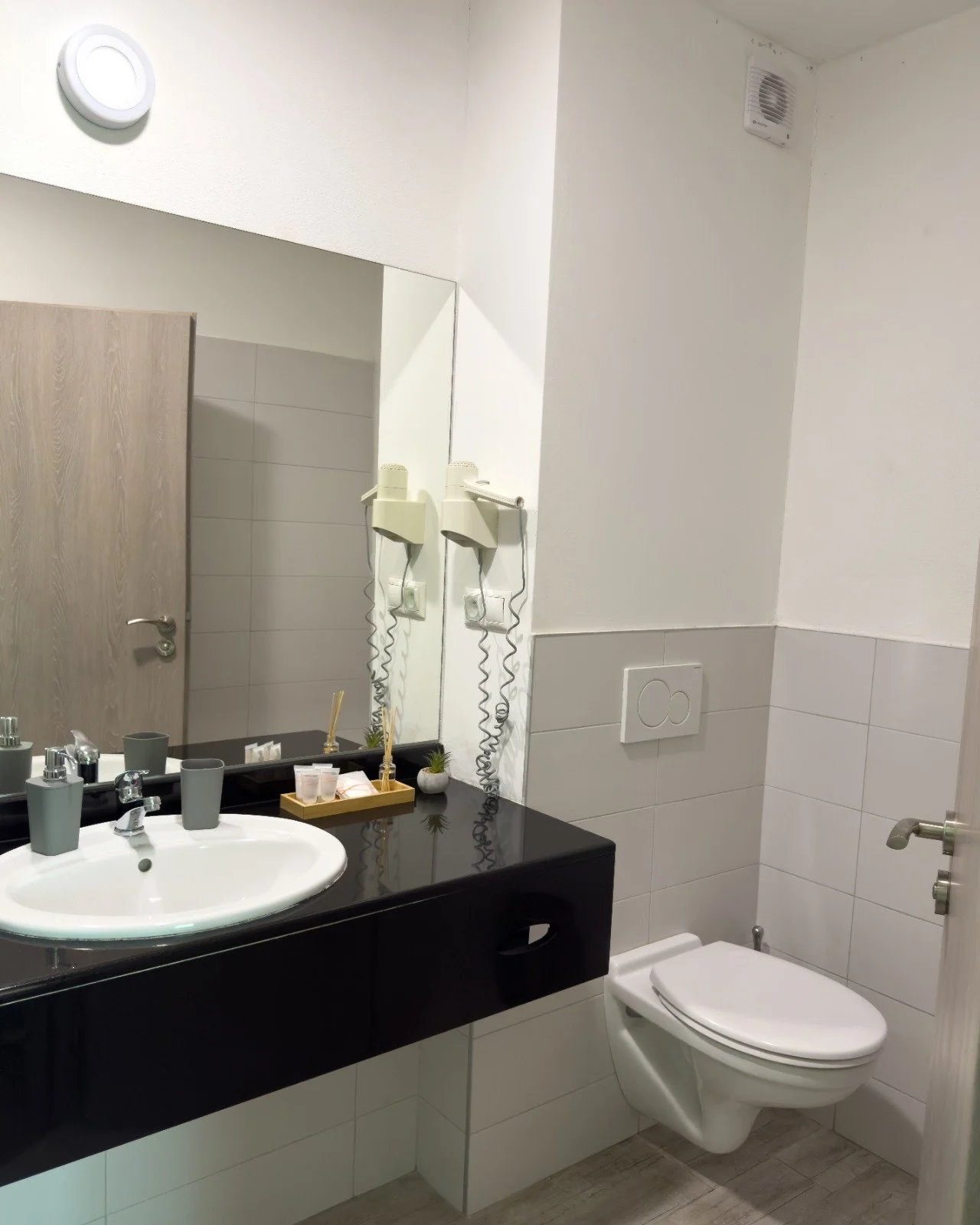 Bathroom with a white toilet and sink, black countertop with toiletries, large mirror, wall-mounted hairdryer, ventilation fan, and light-colored walls and tiles.