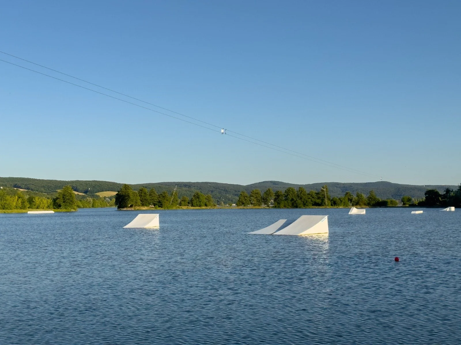 A lake with a series of white ramps or obstacles floating on the water, surrounded by green trees and hills in the background, under a clear blue sky, with a cable car line crossing the sky.
