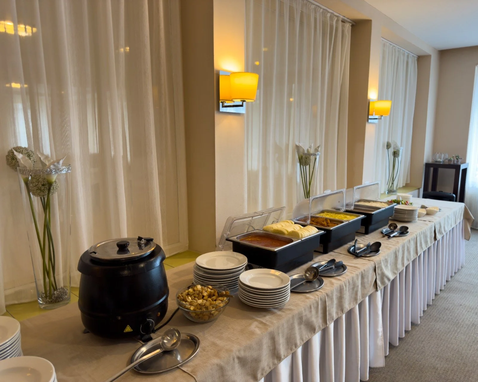Buffet table with various dishes, plates, and utensils in a dining area with cream-colored drapes and wall lamps.