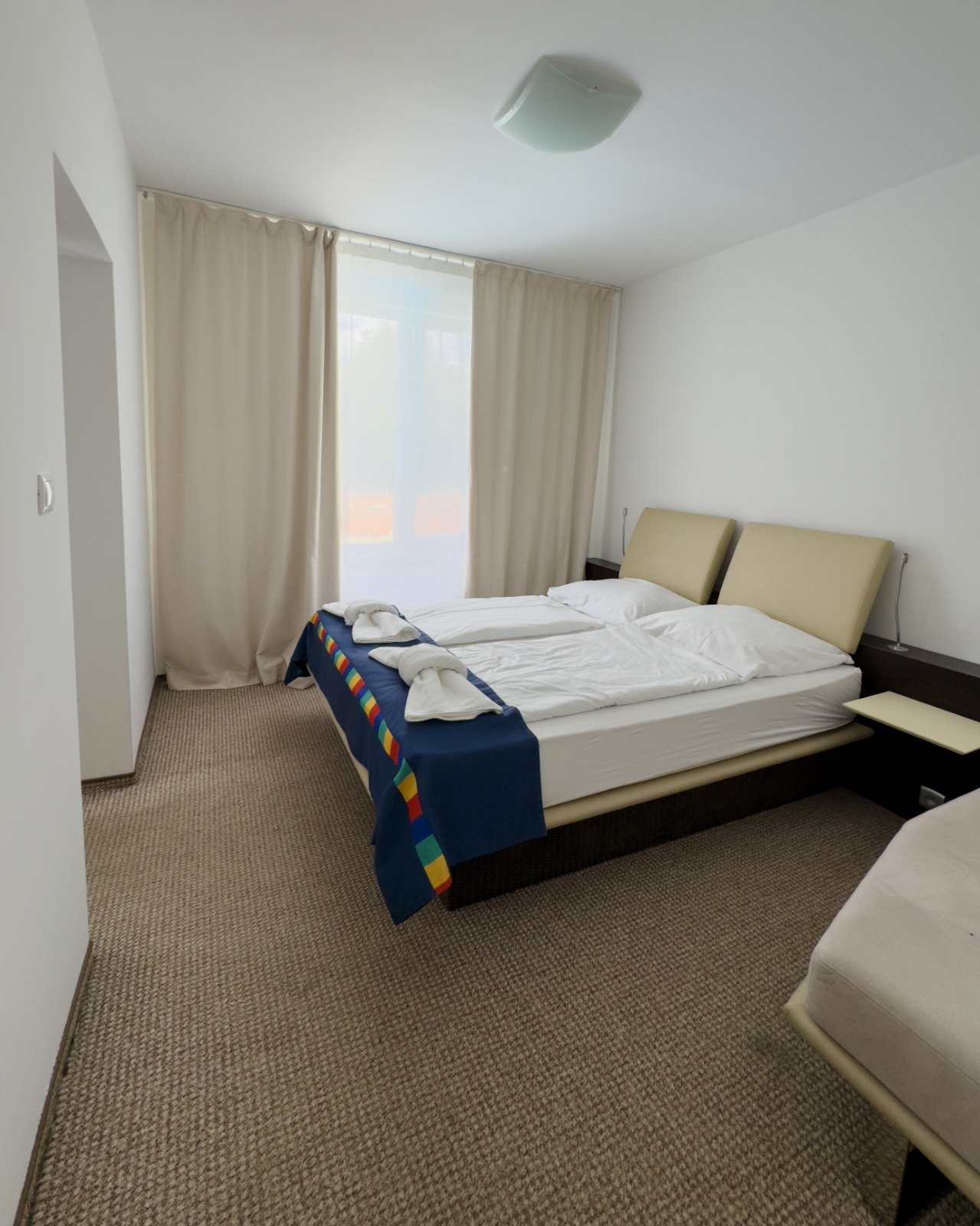 Simple hotel room with two single beds pushed together, beige headboards, white bedding, towels folded on the bed, and curtains covering a window.