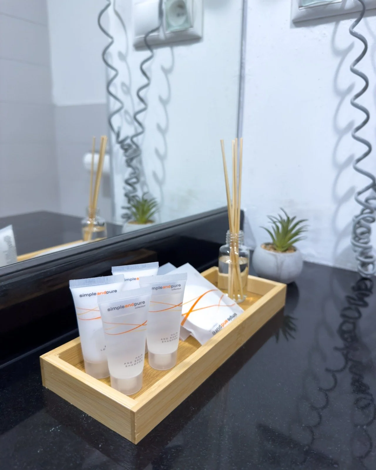 A bathroom countertop with a mirror, small plants in pots, reed diffusers, and toiletries in a wooden tray.