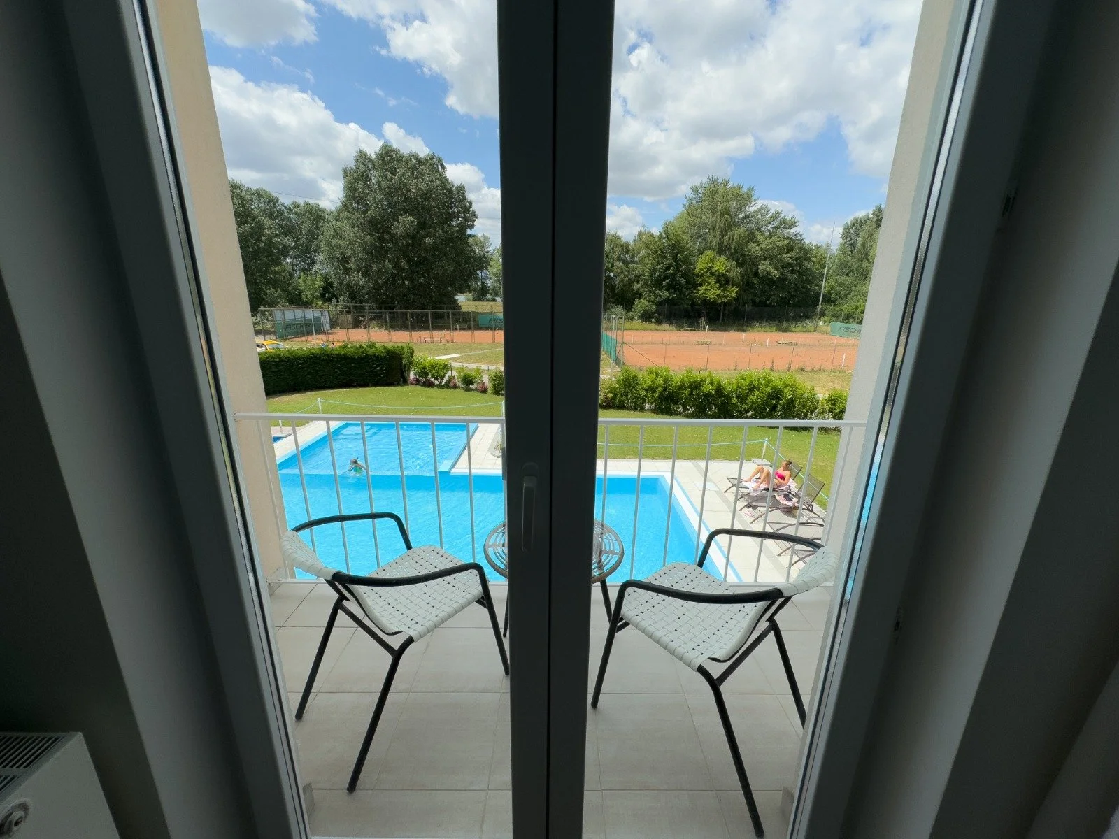View of a swimming pool on a balcony, with two chairs and a small table, overlooking a grassy yard, tennis courts, and trees under a partly cloudy sky.