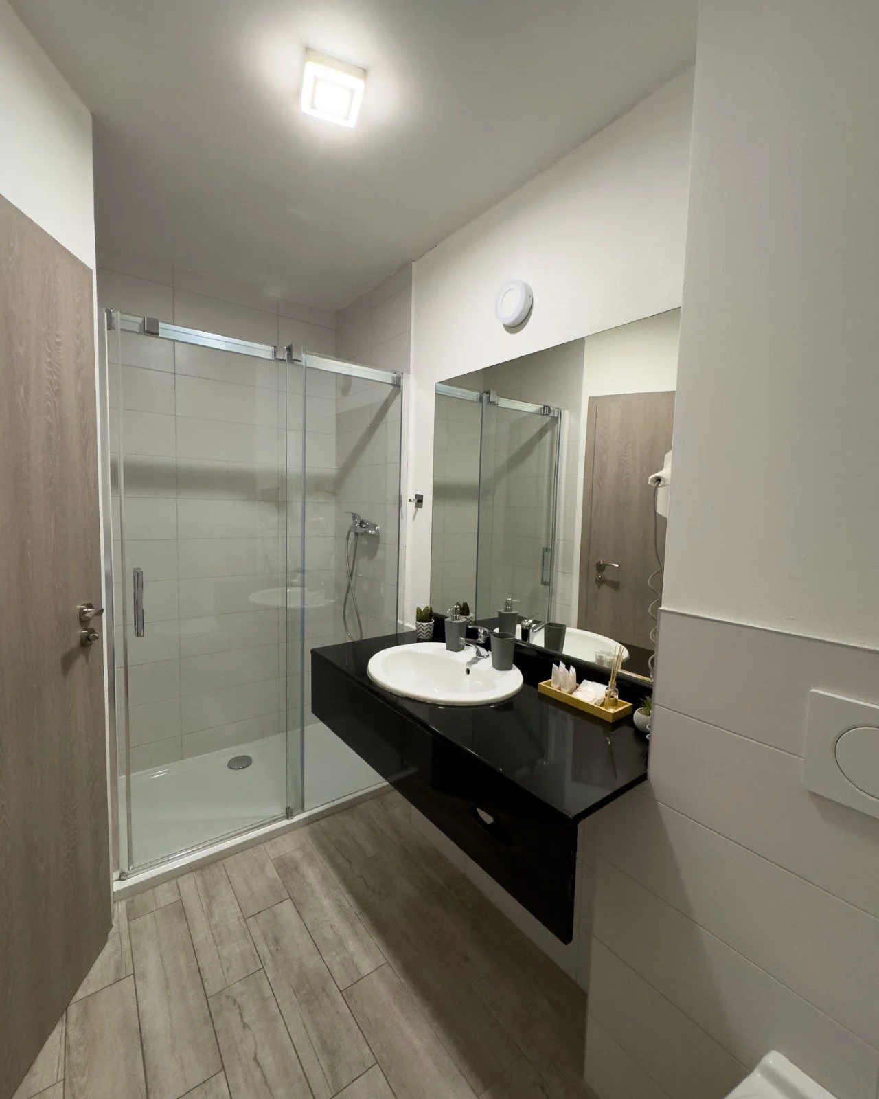 Modern bathroom with a walk-in glass shower, black vanity with a white sink, large mirror, beige wood-look tile flooring, and minimal decor.