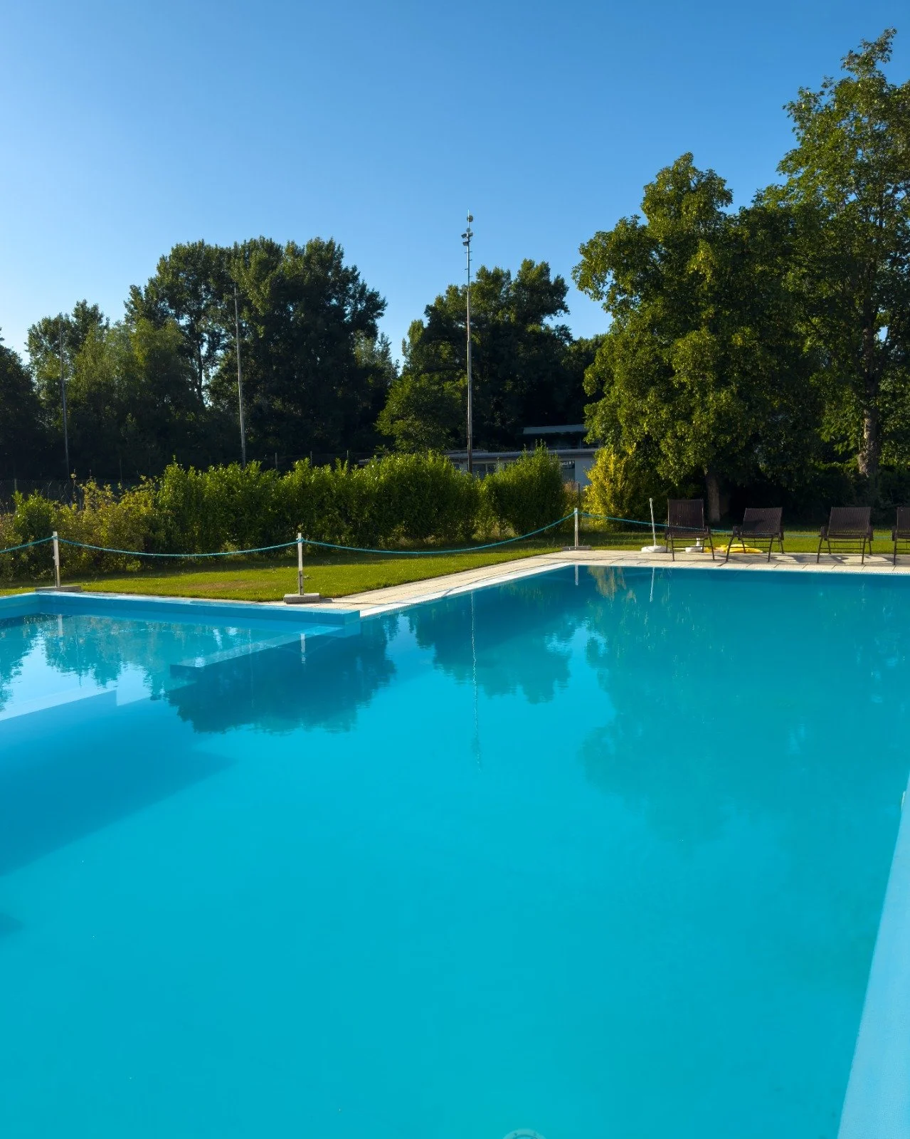 An outdoor swimming pool with clear blue water, surrounded by green grass, trees, and lounge chairs under a bright blue sky.