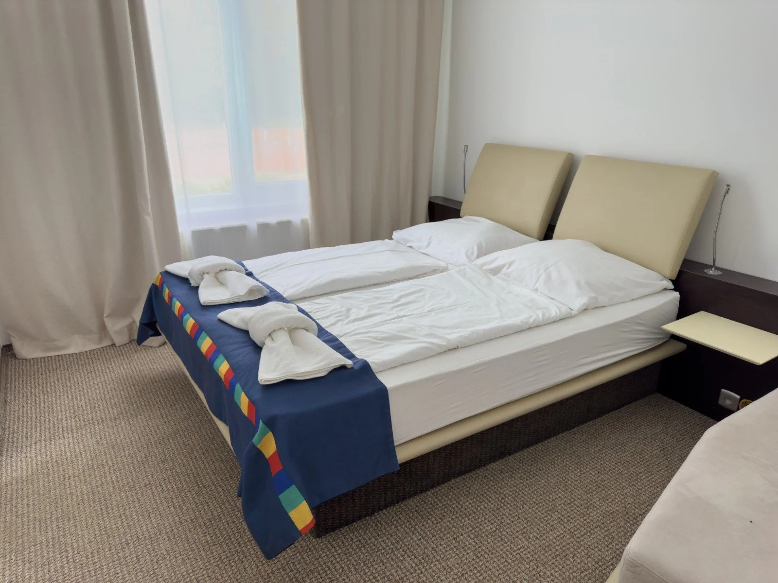 A hotel room with a double bed, two beige headboards, white bedding, two pillows, towel art at the foot of the bed, a nightstand on each side, a window with sheer curtains, and carpeted floor.