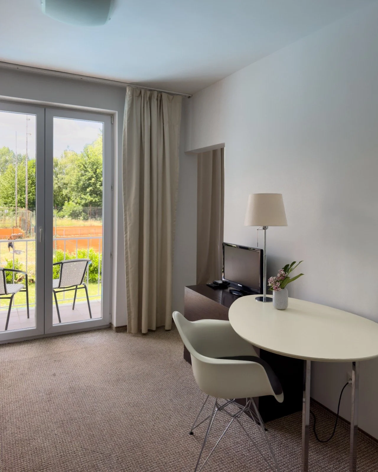 Living room with sliding glass door leading to outdoor balcony, beige curtains, white wall, small flat-screen TV on wooden stand, white table with chair, potted plant, and floor lamp.