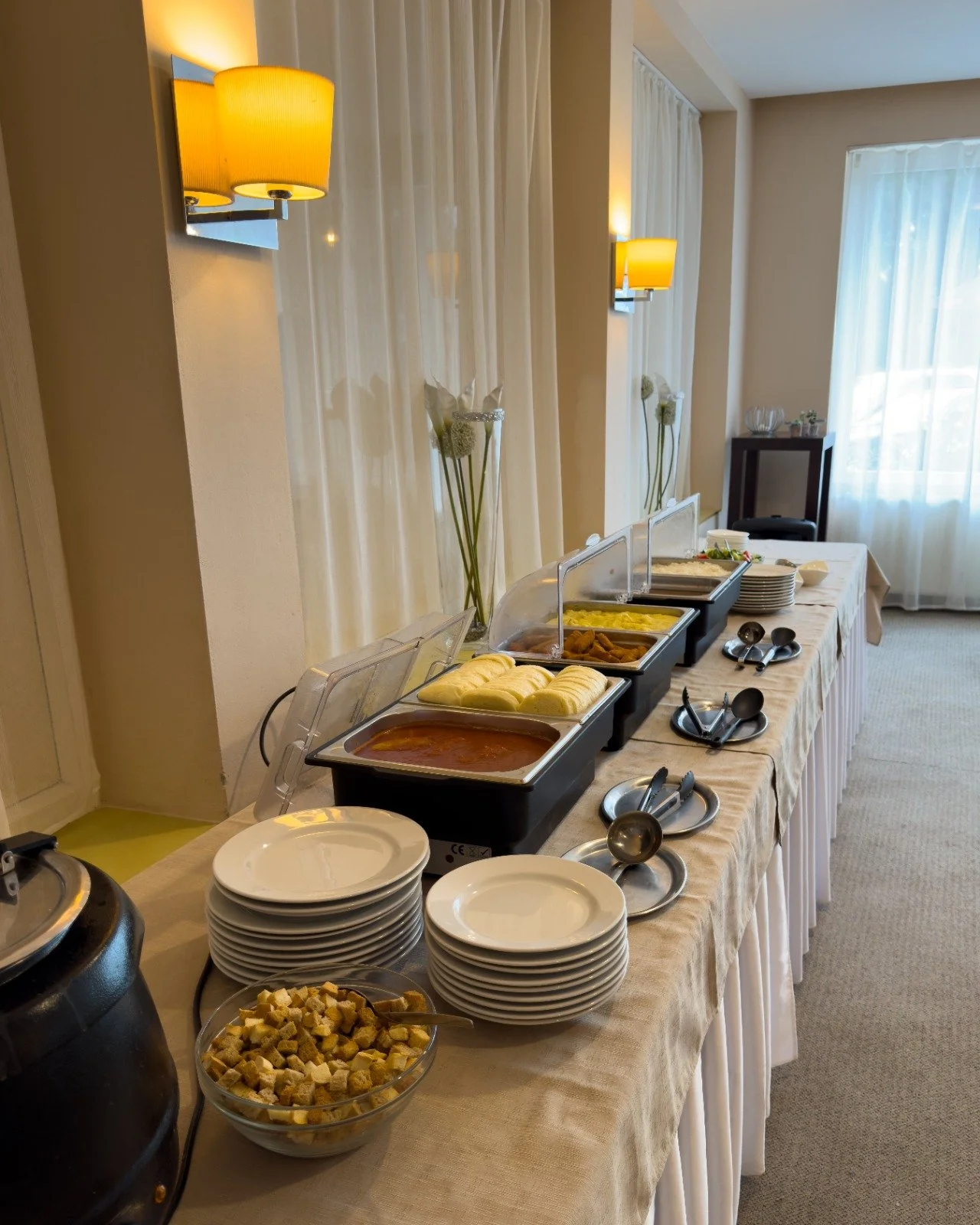 Buffet table with asorted breakfast foods including scrambled eggs, bacon, bread rolls, cereal, and condiments, set in a hotel dining area with curtains and wall lamps.