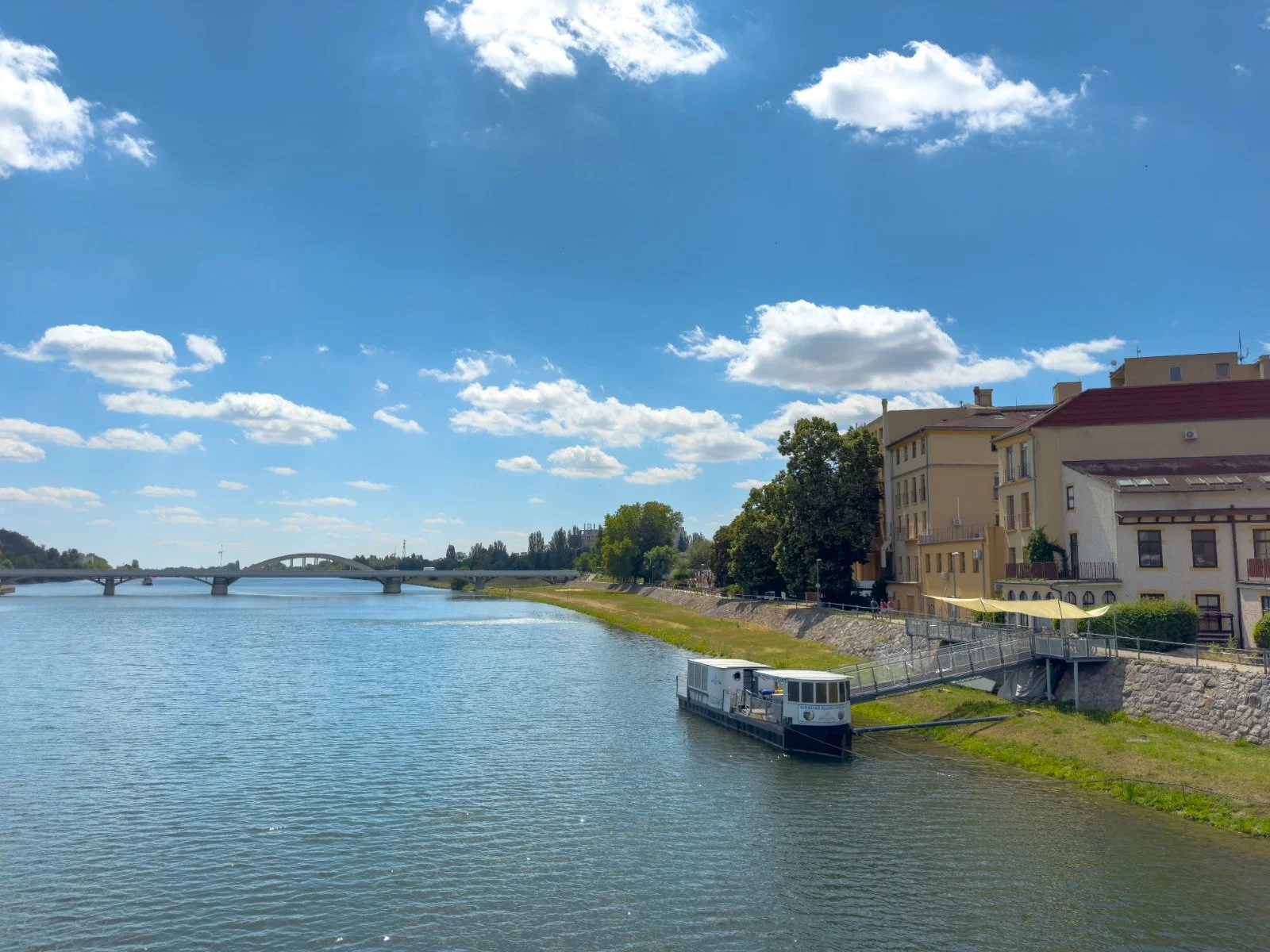 A scenic view of a river with a boat docked along the bank, a bridge in the background, and colorful buildings with yellow awnings on the right side under a partly cloudy sky.