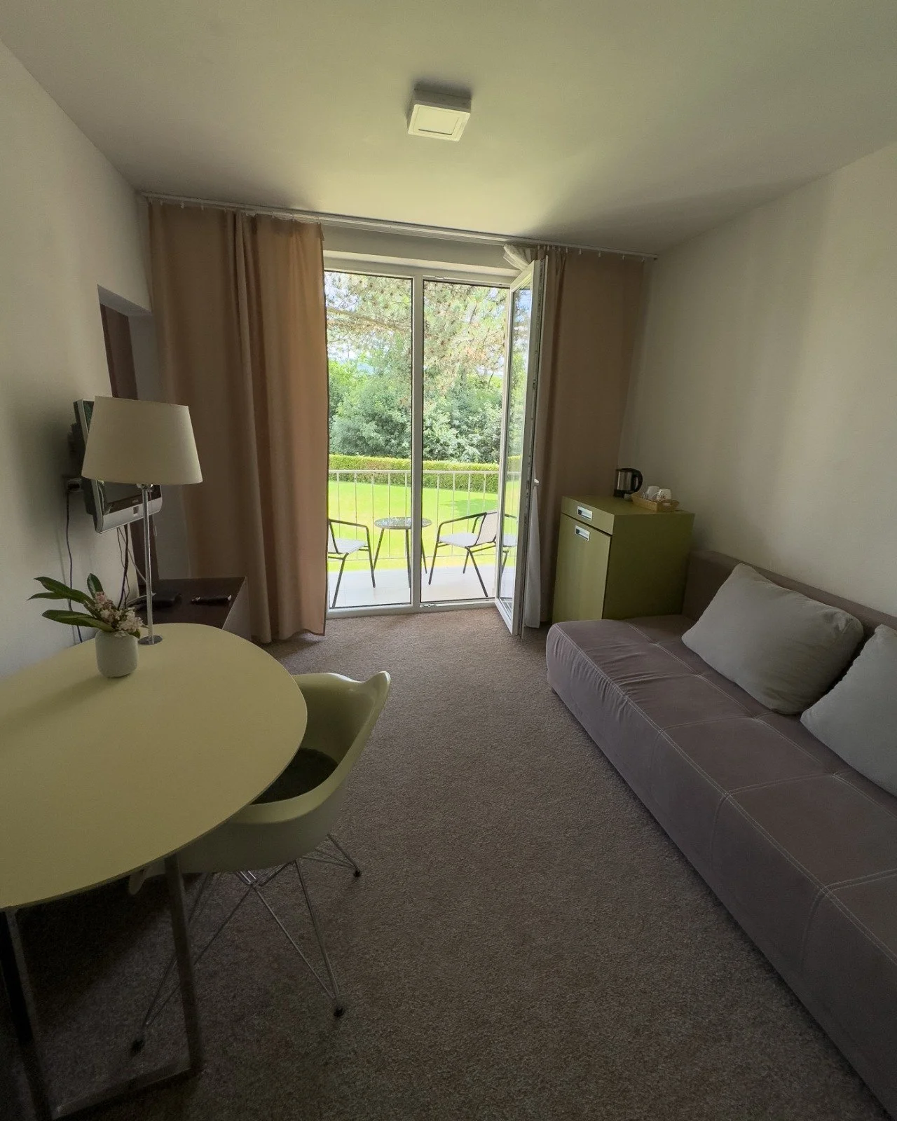A hotel room with a sliding glass door leading to a balcony with outdoor chairs and a view of green trees and grass.