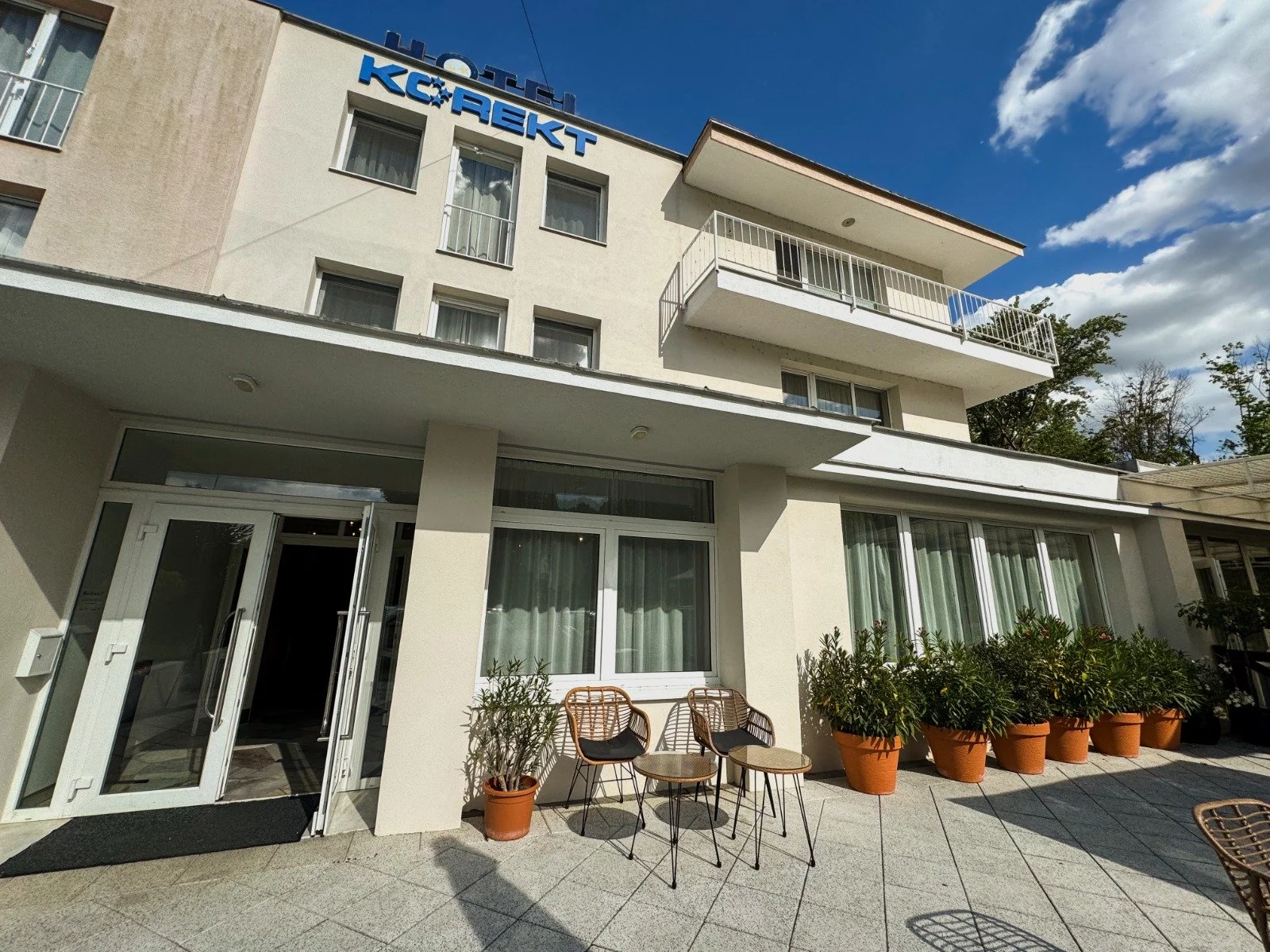 Hotel Krek T building with outdoor patio area, potted plants, chairs, and glass door entrance, under a partly cloudy sky.