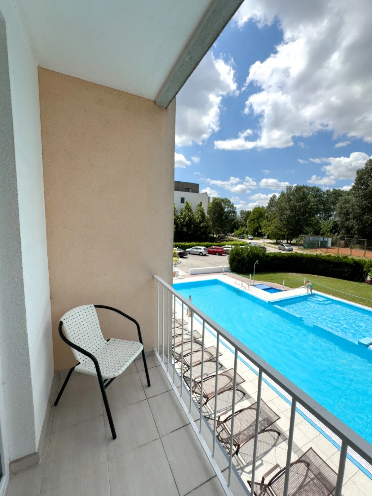 View from a balcony overlooking a swimming pool with clear blue water, several chairs around it, a person swimming in the pool, trees, a parking lot with cars, and a partly cloudy sky.