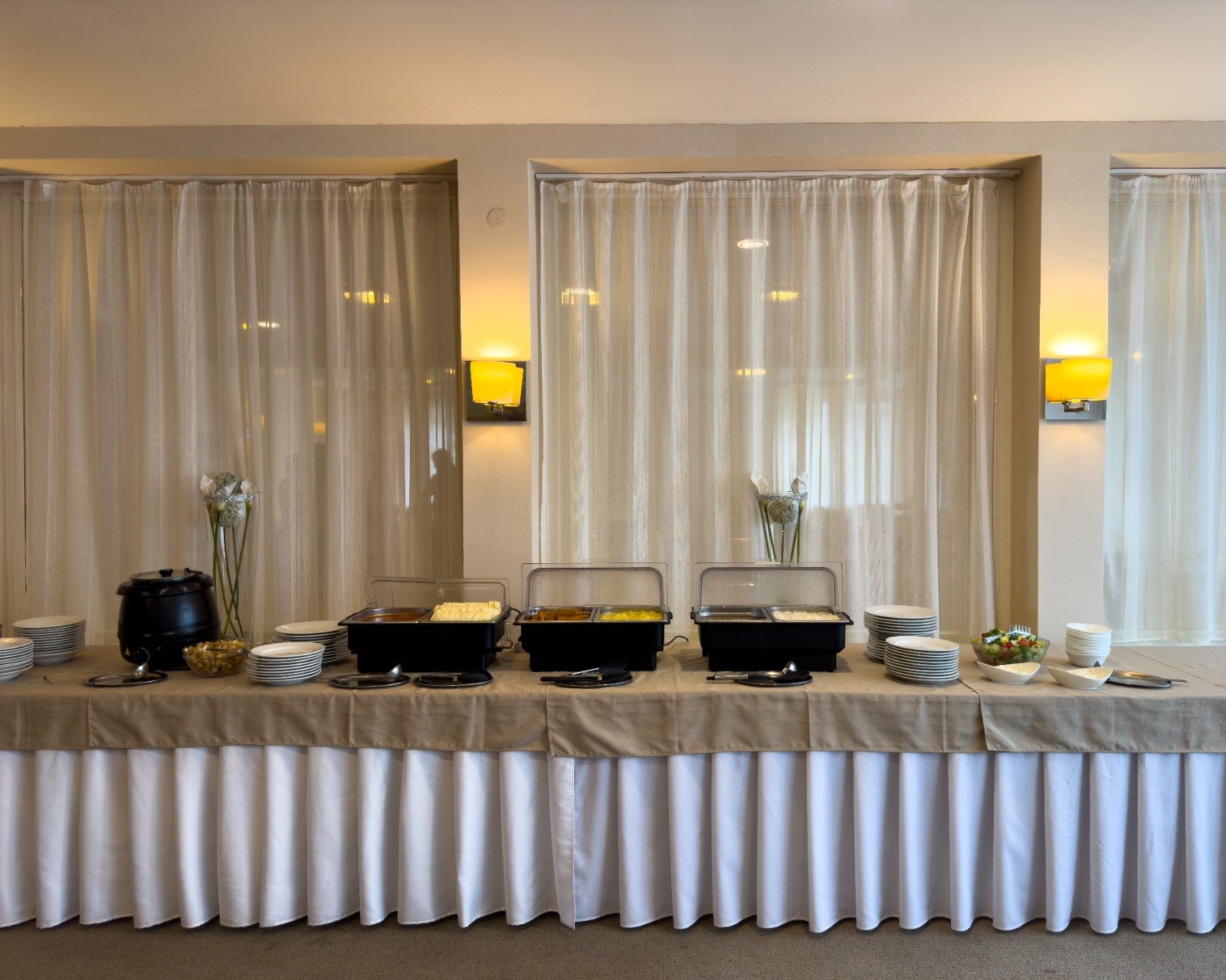 Buffet table with chafing dishes, bowls of salad and condiments, stacks of plates and bowls, and decorative vases with flowers, set against a backdrop of beige curtains and yellow wall sconces.
