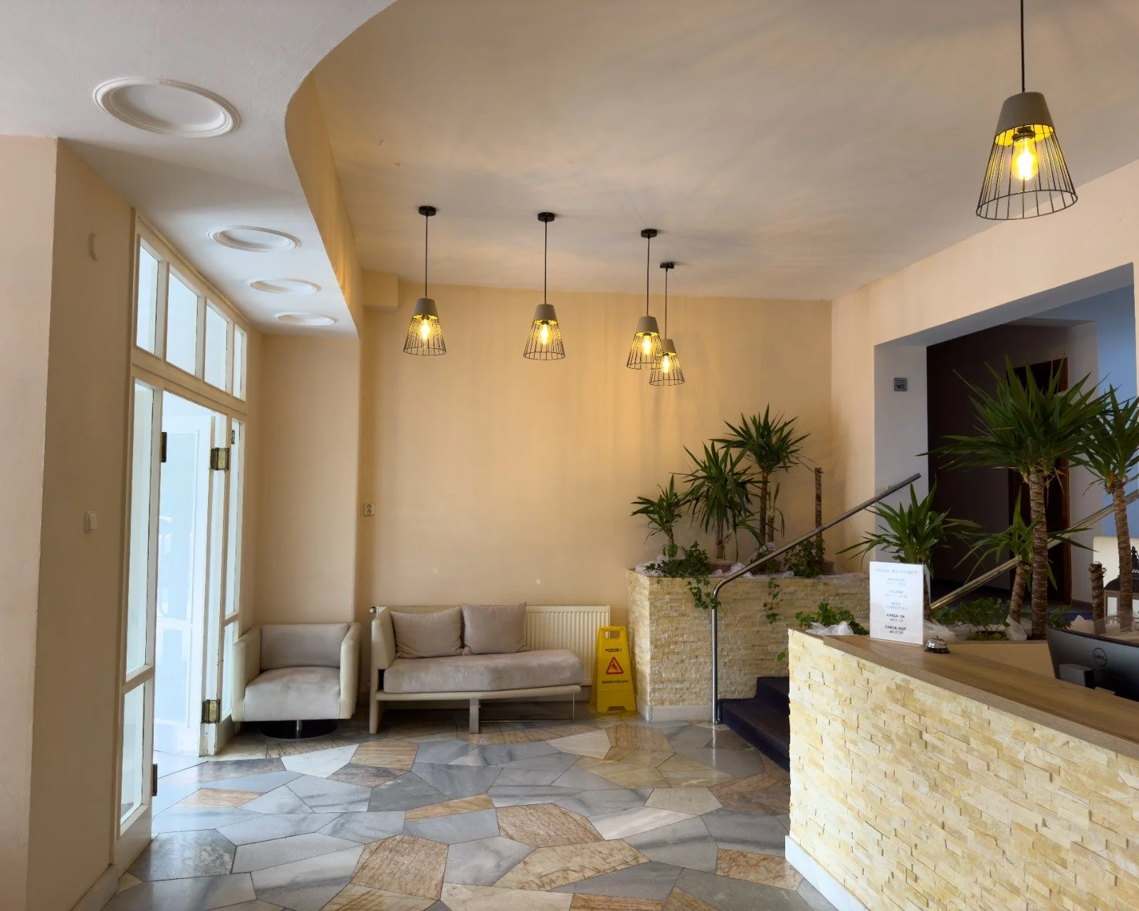 Hotel lobby with a reception desk, potted plants, a seating area with two beige chairs, and pendant lights hanging from the ceiling.