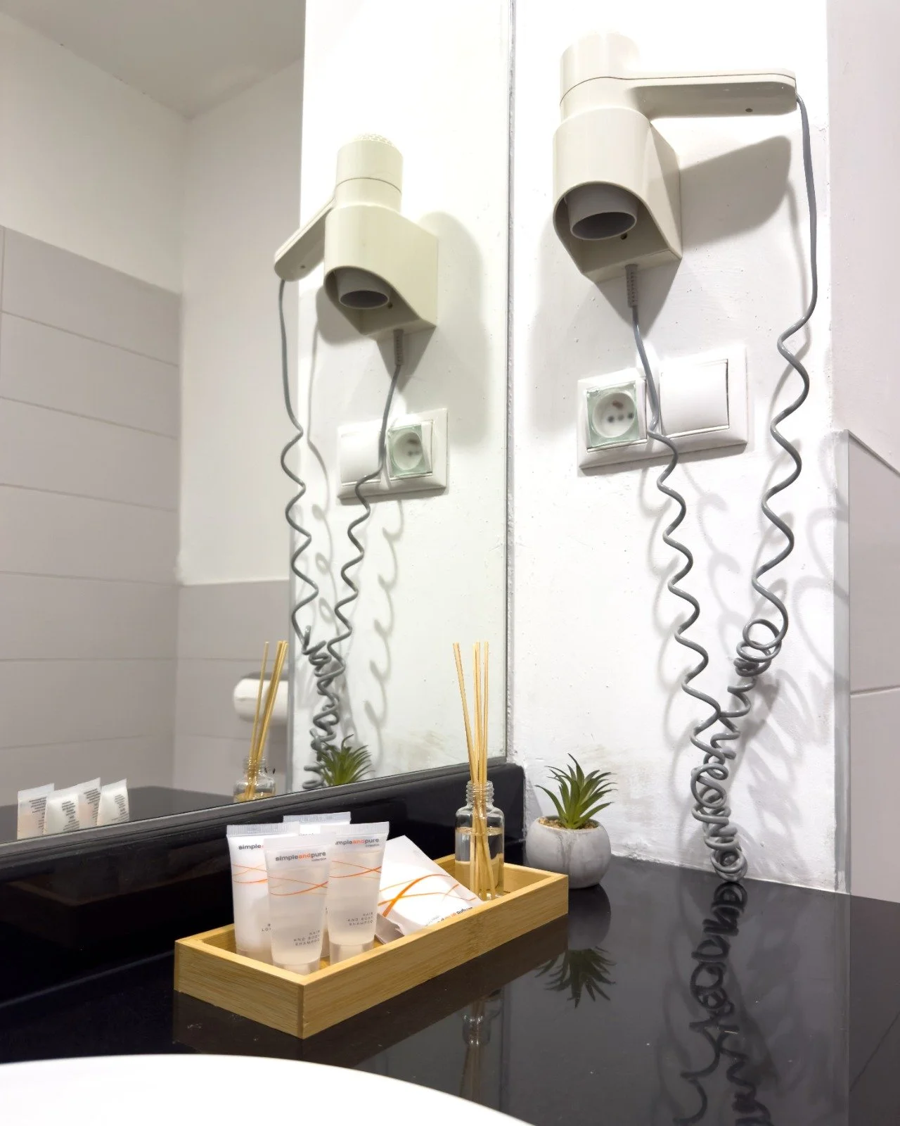 A bathroom with a large mirror, a wall-mounted white hair dryer, a power outlet, a black countertop with a wooden tray holding lotions and reed diffusers, and small potted plants.