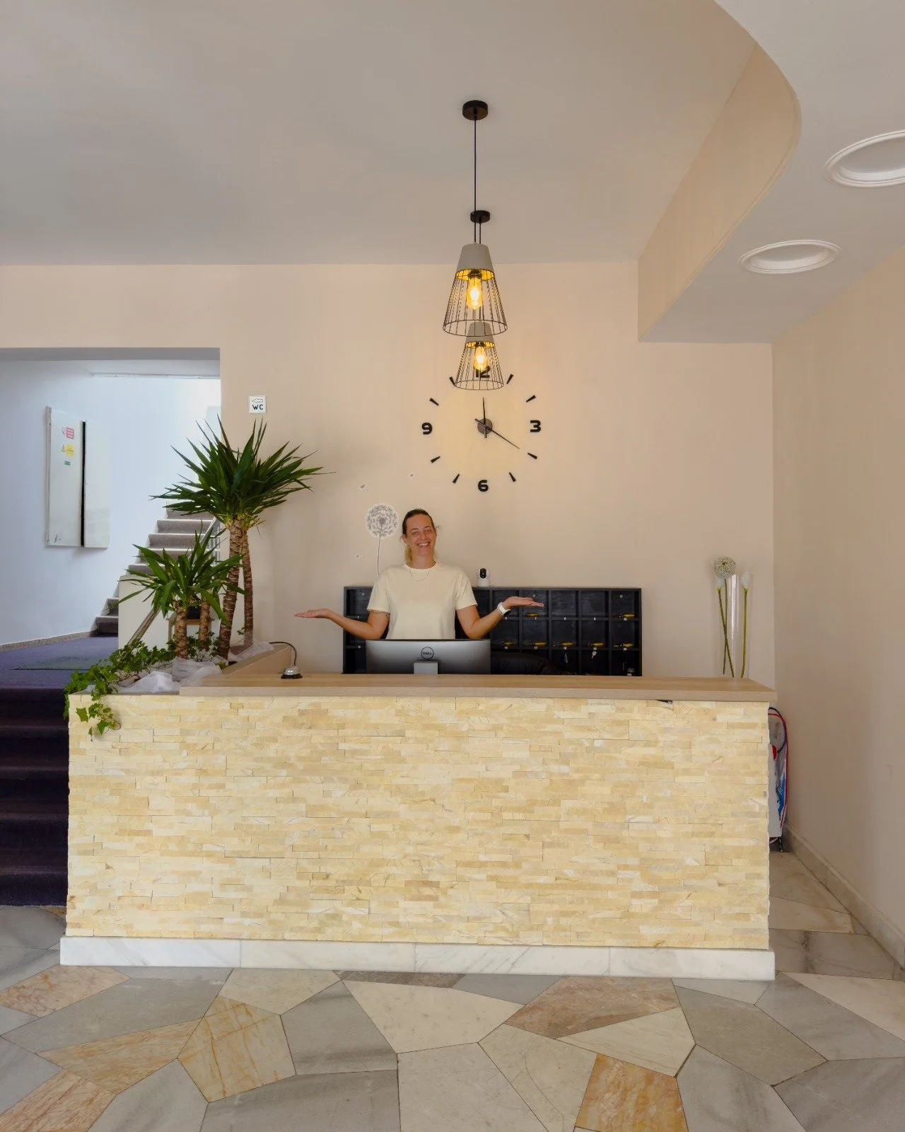A woman standing behind a reception desk with a computer, smiling with arms outstretched. Behind her is a large wall clock, and to her left are two potted plants. The reception area has a modern design with a light-colored stone facade and geometric 