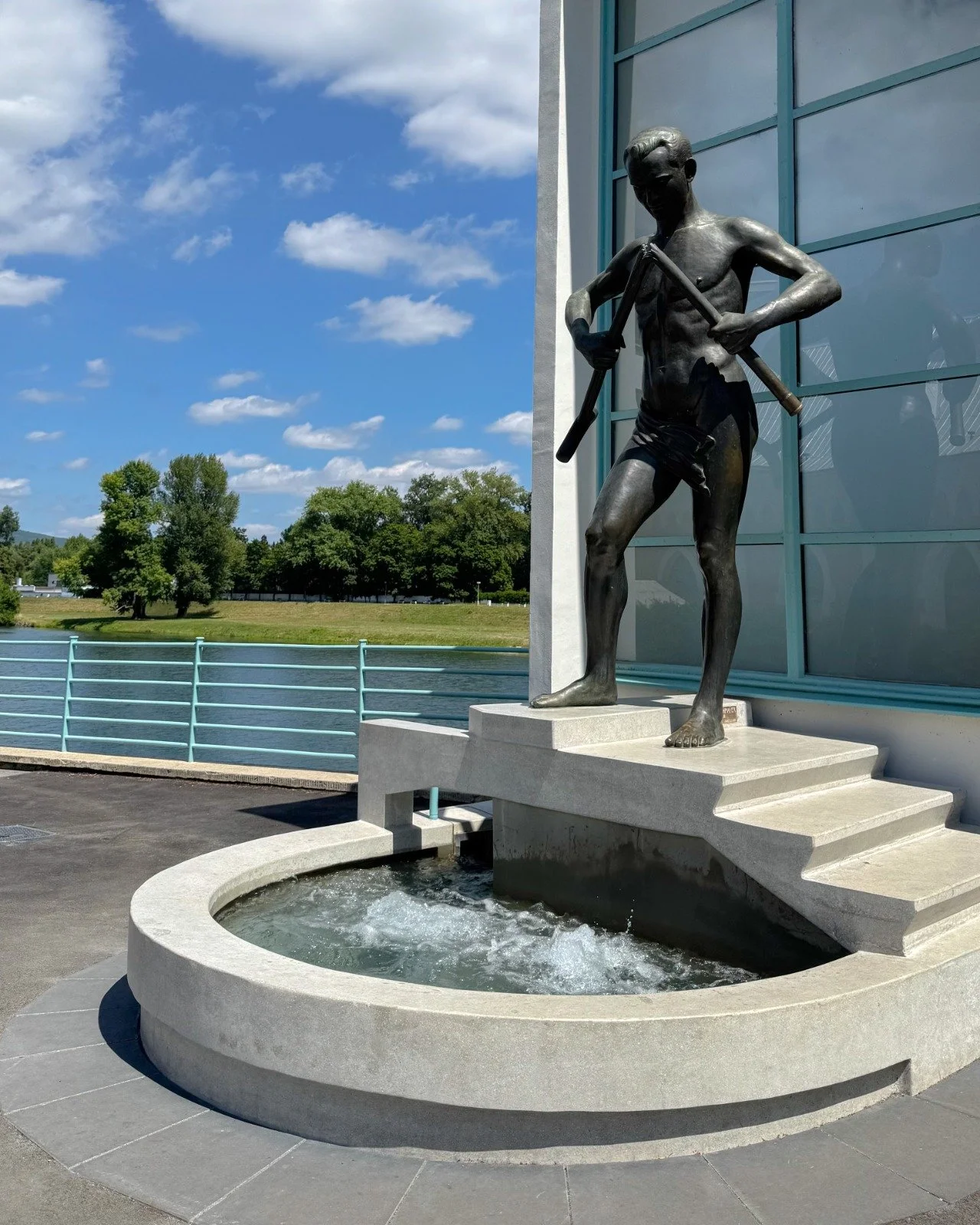 A bronze statue of a young boy standing on a pedestal with a small water fountain below. The boy holds a stick across his shoulders and is shirtless, wearing shorts. The statue is located outdoors next to a modern building with large windows, with tr