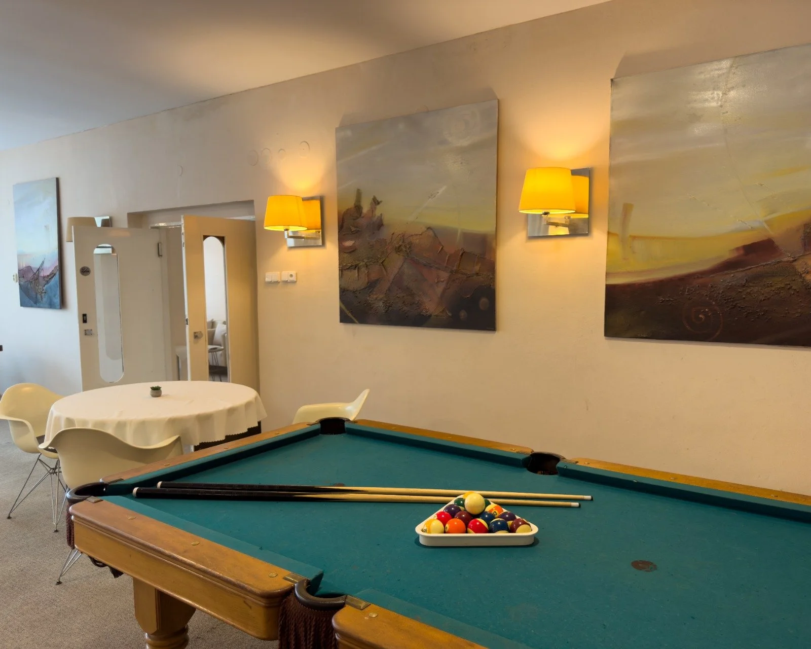 A pool table with pool balls arranged at the end, two pool cues resting on the edge, inside a room with white walls, artwork, wall lamps, a round table with a small plant, and chairs.