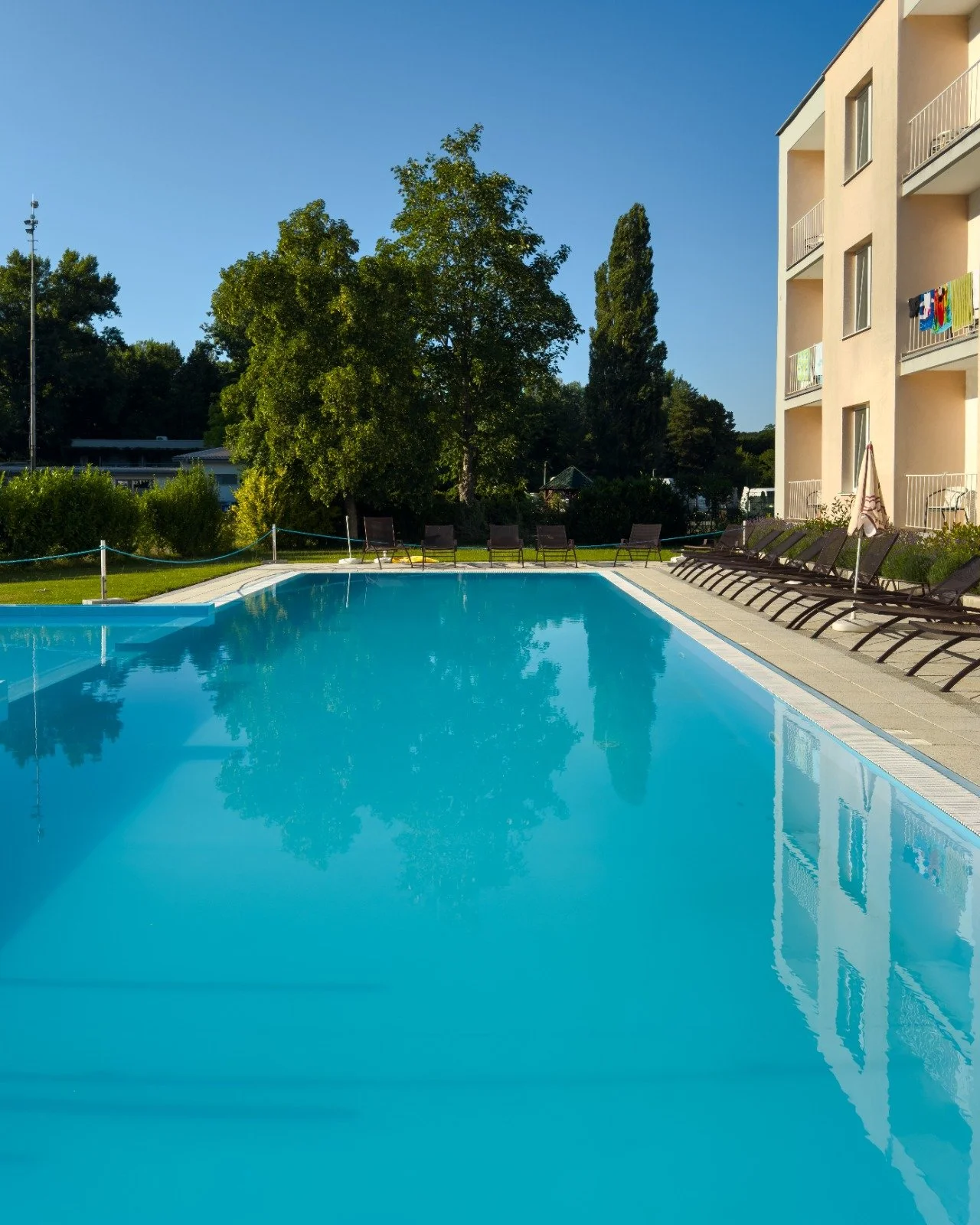 Swimming pool at an apartment complex with lounge chairs, a building with balconies, and trees in the background under a clear blue sky.