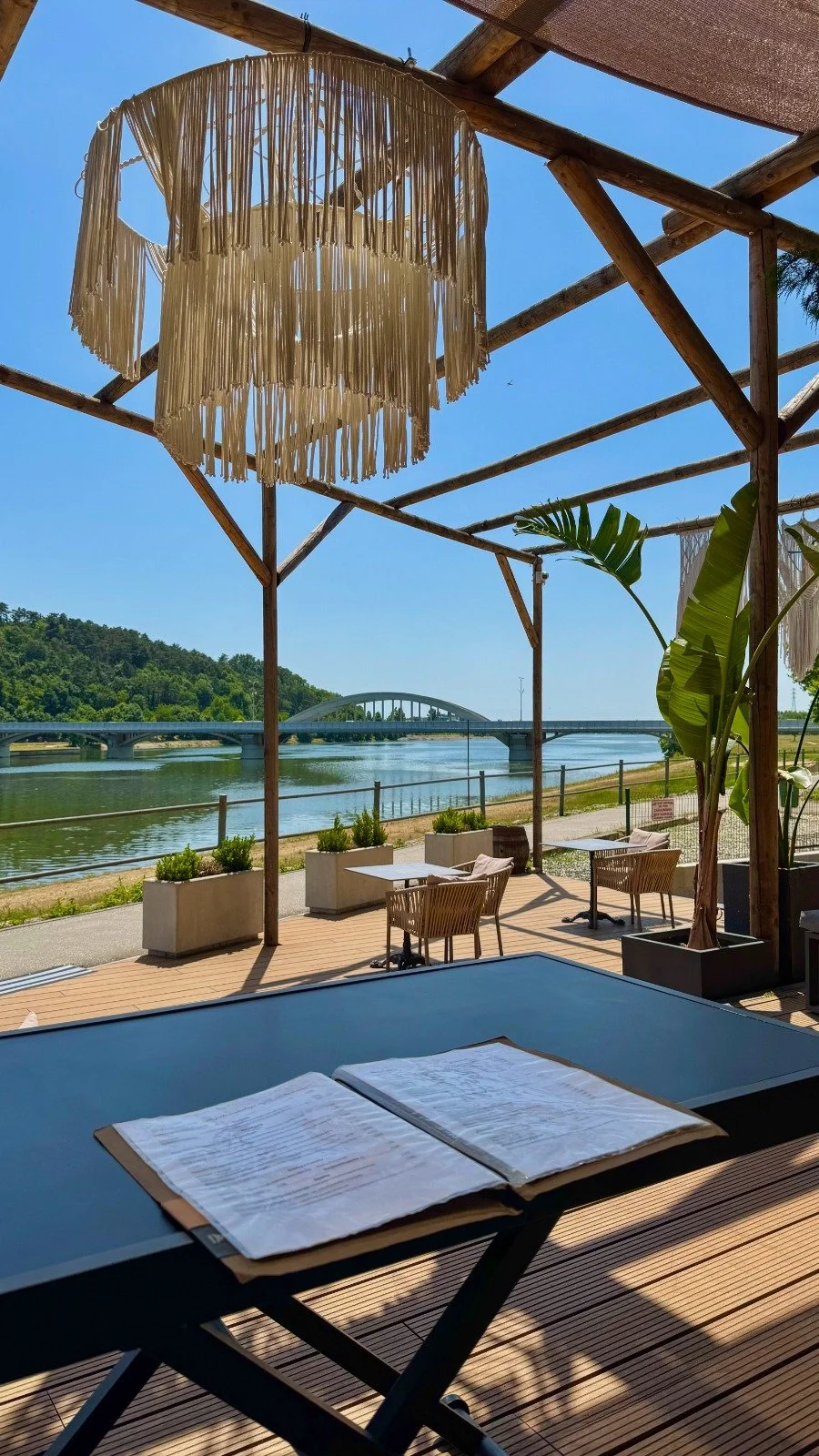 Outdoor seating area overlooking a river with a bridge in the distance, under a wooden pergola with a hanging fringed lamp and potted plants.