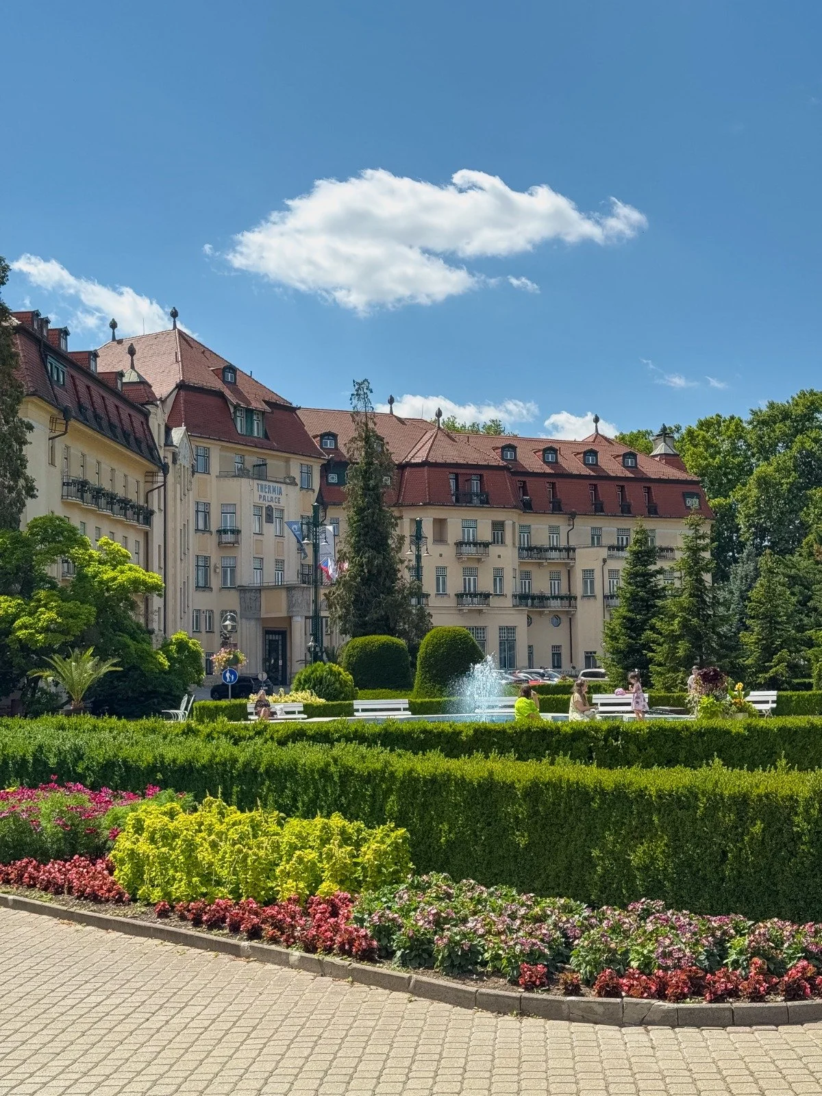 A sunny day in a park with a fountain, surrounded by colorful flowers and neatly trimmed bushes, with a large hotel or apartment building in the background and a bright blue sky with a few clouds.