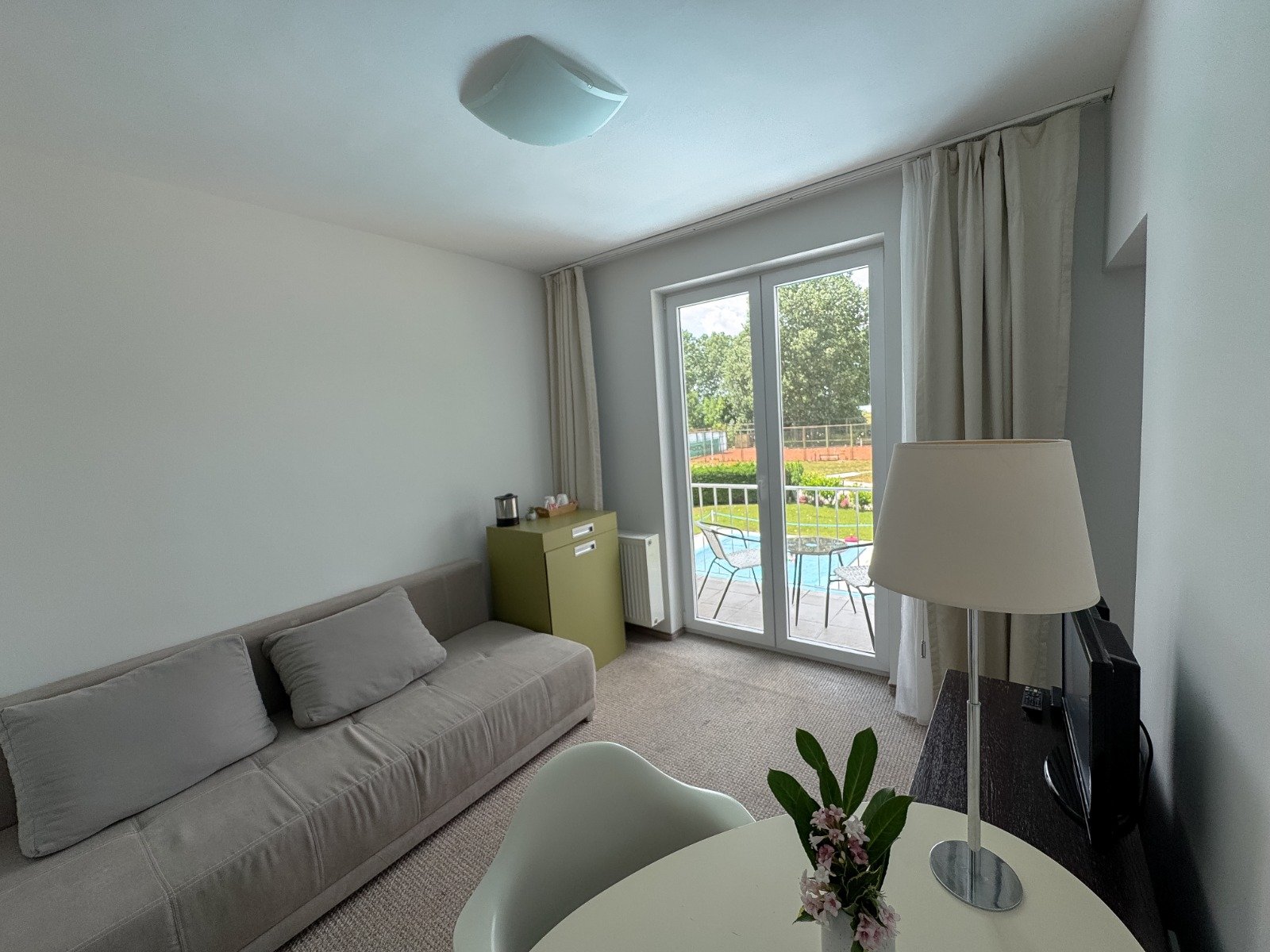 Living room with a gray sofa, a small green cabinet, a floor lamp, a television, and a round dining table with a vase of flowers. Large glass door leading to a balcony with outdoor furniture, overlooking green trees and a sports field.