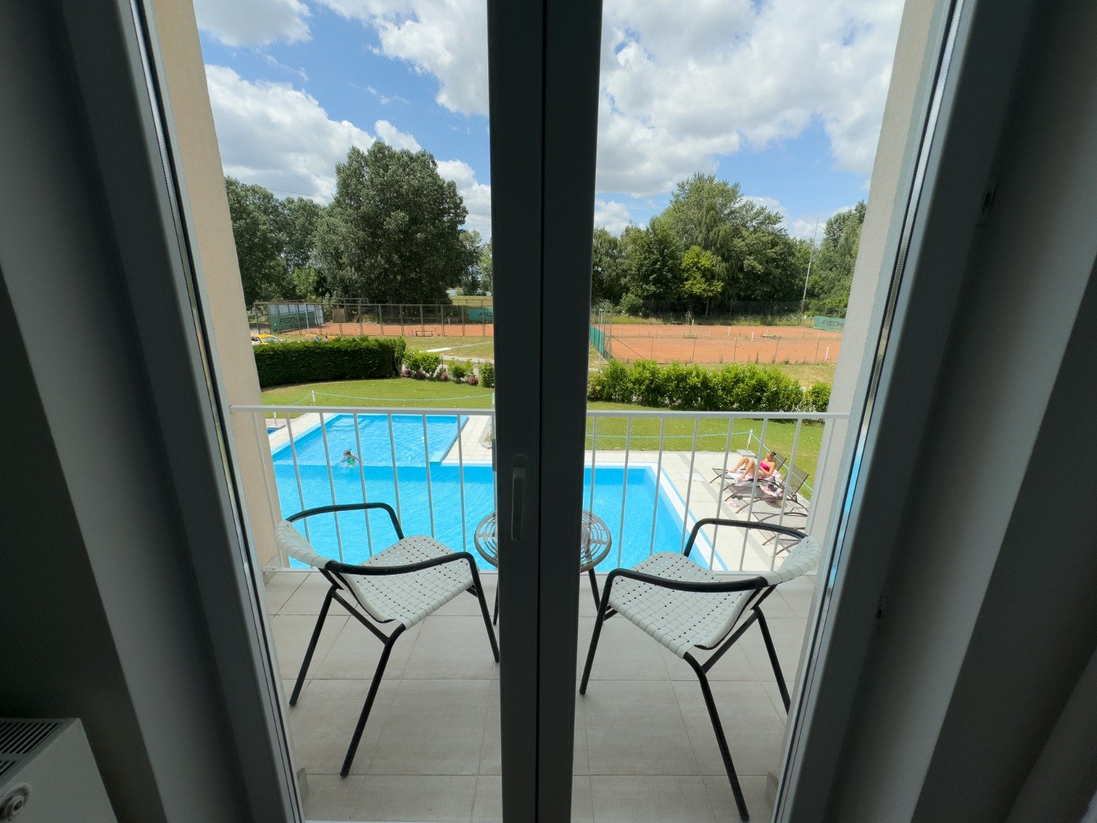View from a balcony looking out onto a swimming pool, a person relaxing on a lounge chair, tennis courts, trees, and a partly cloudy sky.