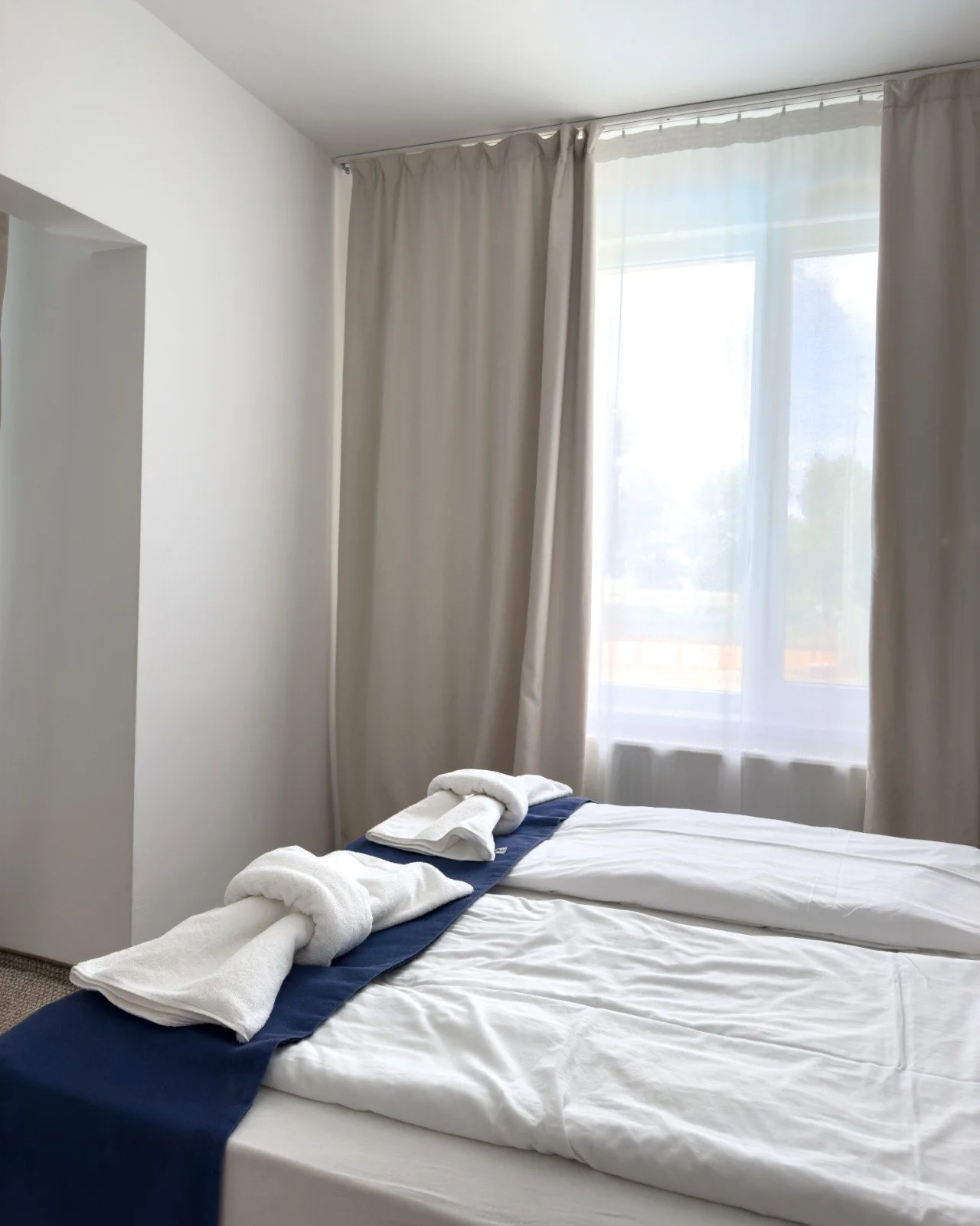 A neatly made hotel bed with white sheets, a dark blue runner, and two rolled towels shaped like swans placed at the foot of the bed. A towel is folded into a swan shape on the bed. There is a window with beige curtains and white sheer curtains that 