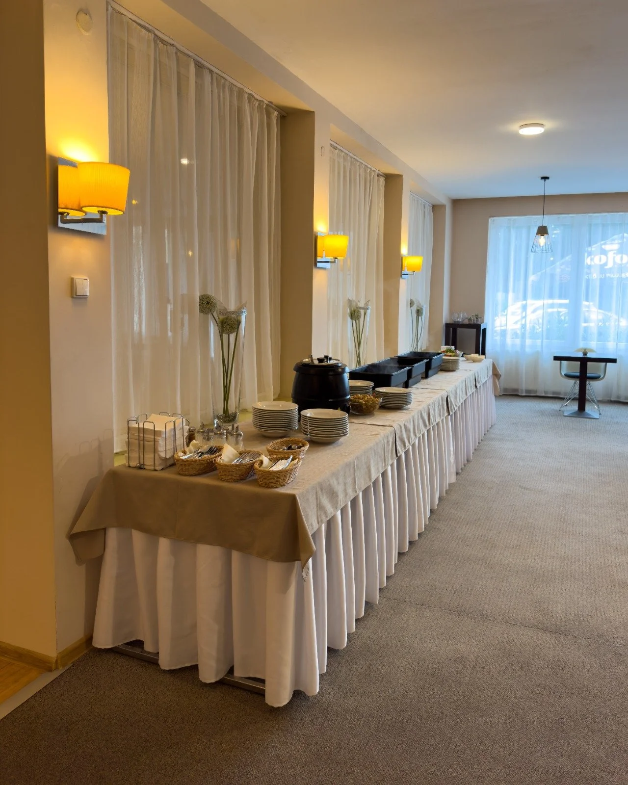 A buffet setup in a hotel breakfast area with a long table covered in a beige tablecloth, holding plates, bowls, a coffee urn, and serving dishes. Behind the table are sheer curtains, decorative vases with flowers, and wall-mounted lamps. In the back