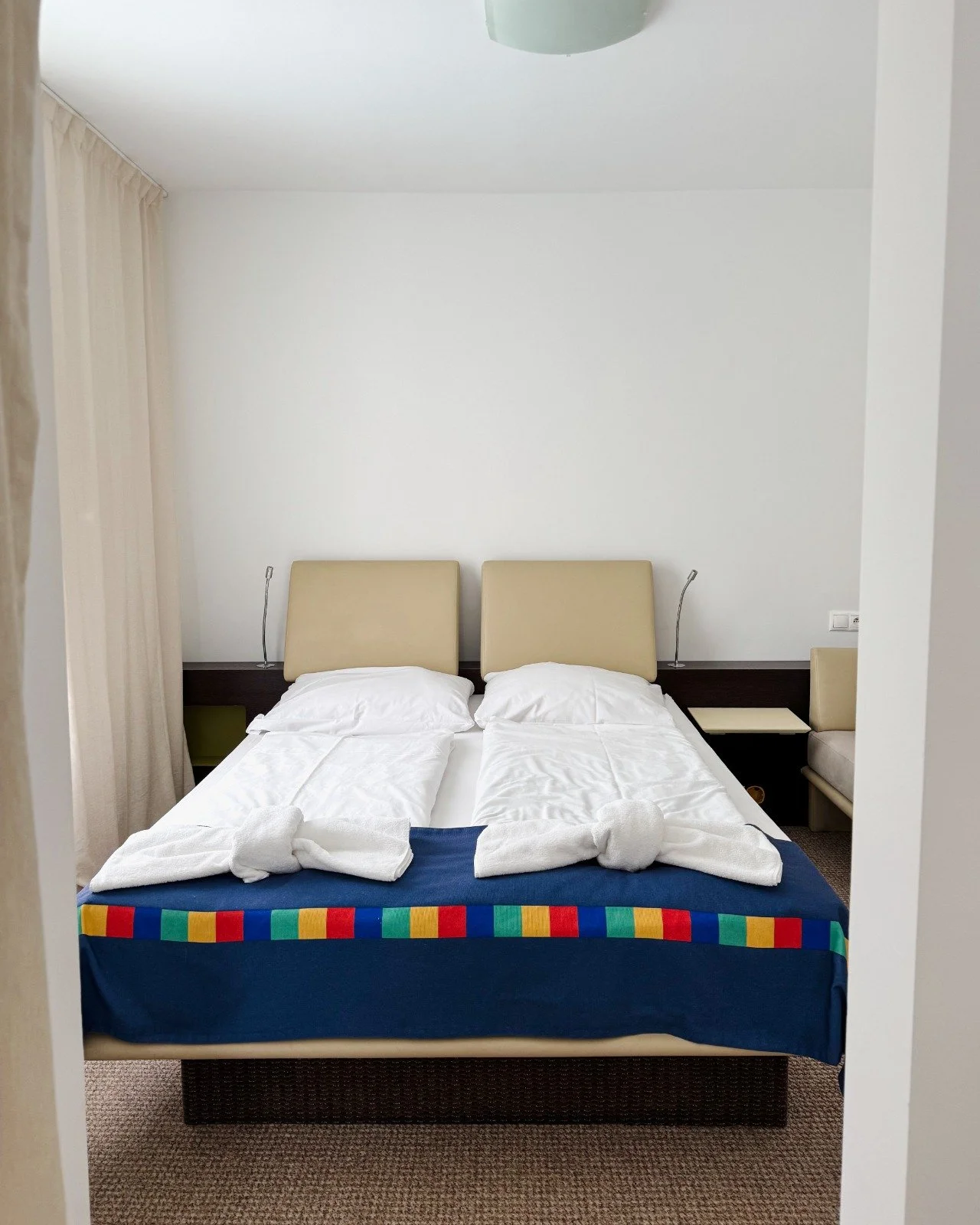 A neatly made hotel bed with two pillows, towel swans, and a colorful bed runner in a minimalist room with beige headboards and side tables.