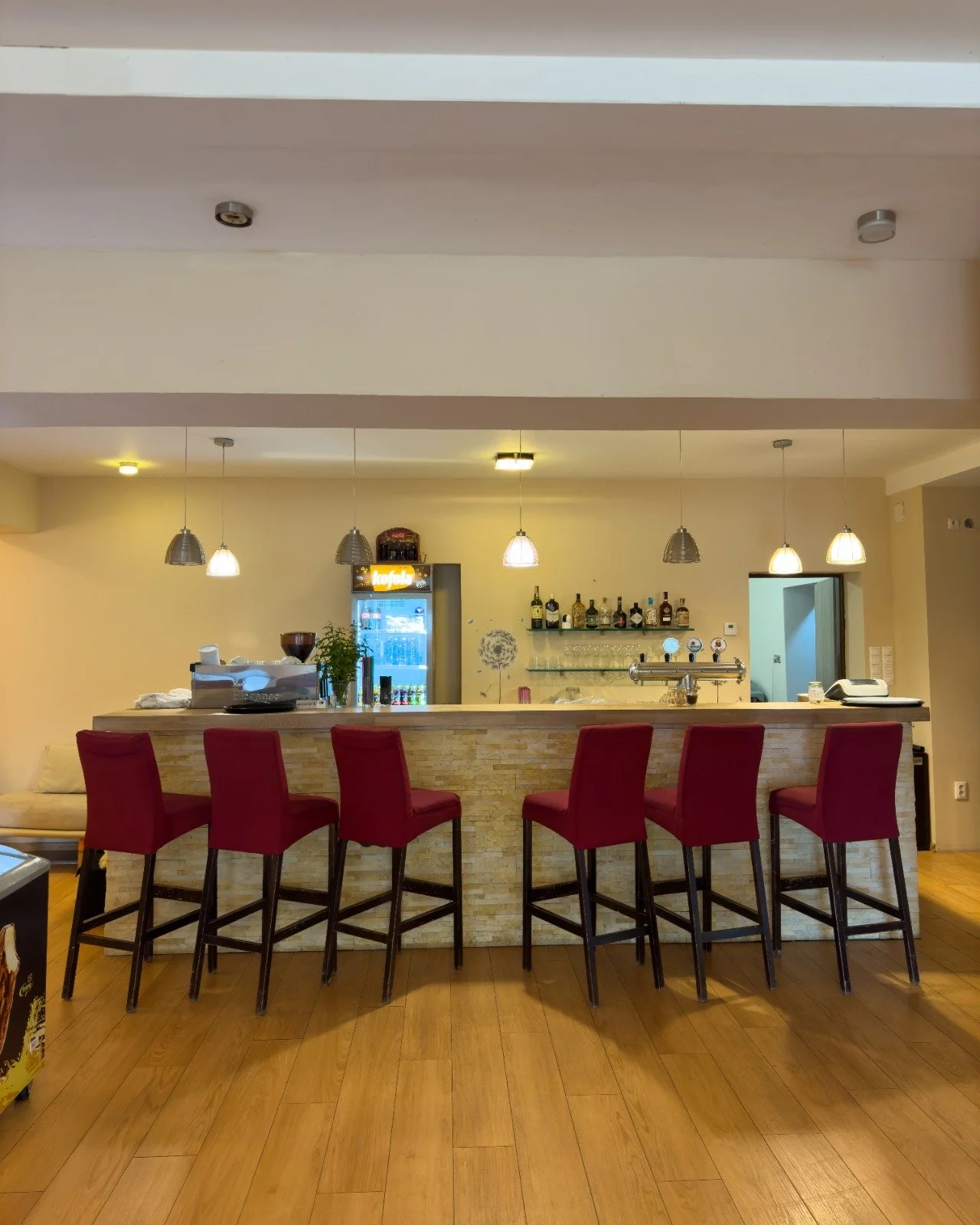A bar with a row of six red chairs at a counter, with espresso machines, liquor bottles, a small fridge, and pendant lights hanging above.