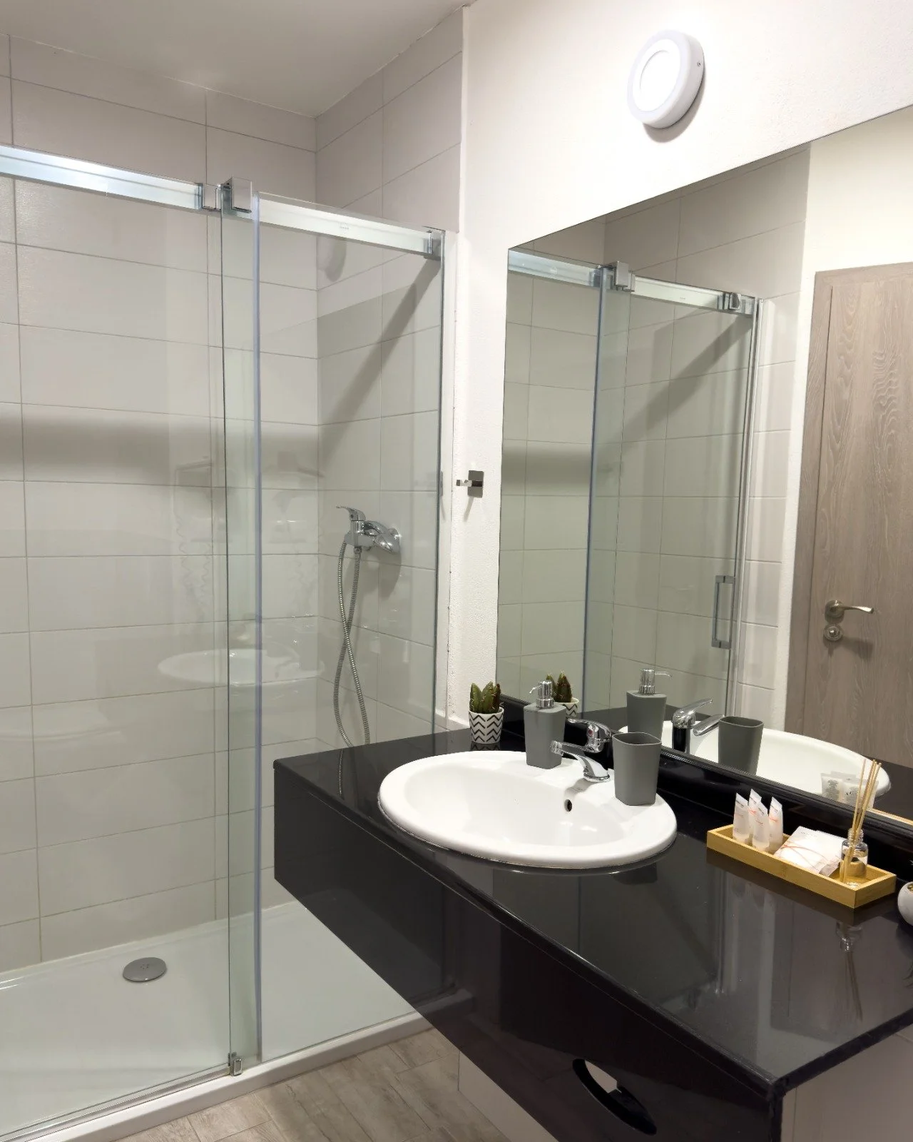 Modern bathroom with a glass shower enclosure, a black vanity with a white sink, and a large mirror. The vanity has gray cups, a soap dispenser, and small decorative items on a wooden tray. There are small potted plants on the counter and a wooden do