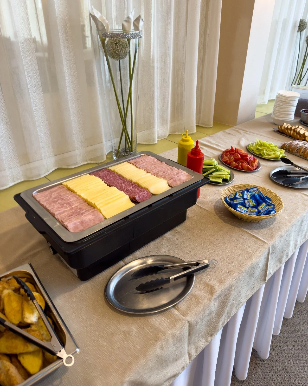 Buffet table with sliced meats, cheese, vegetables, and condiments, along with plates and utensils, set against a backdrop of curtains and flowers.
