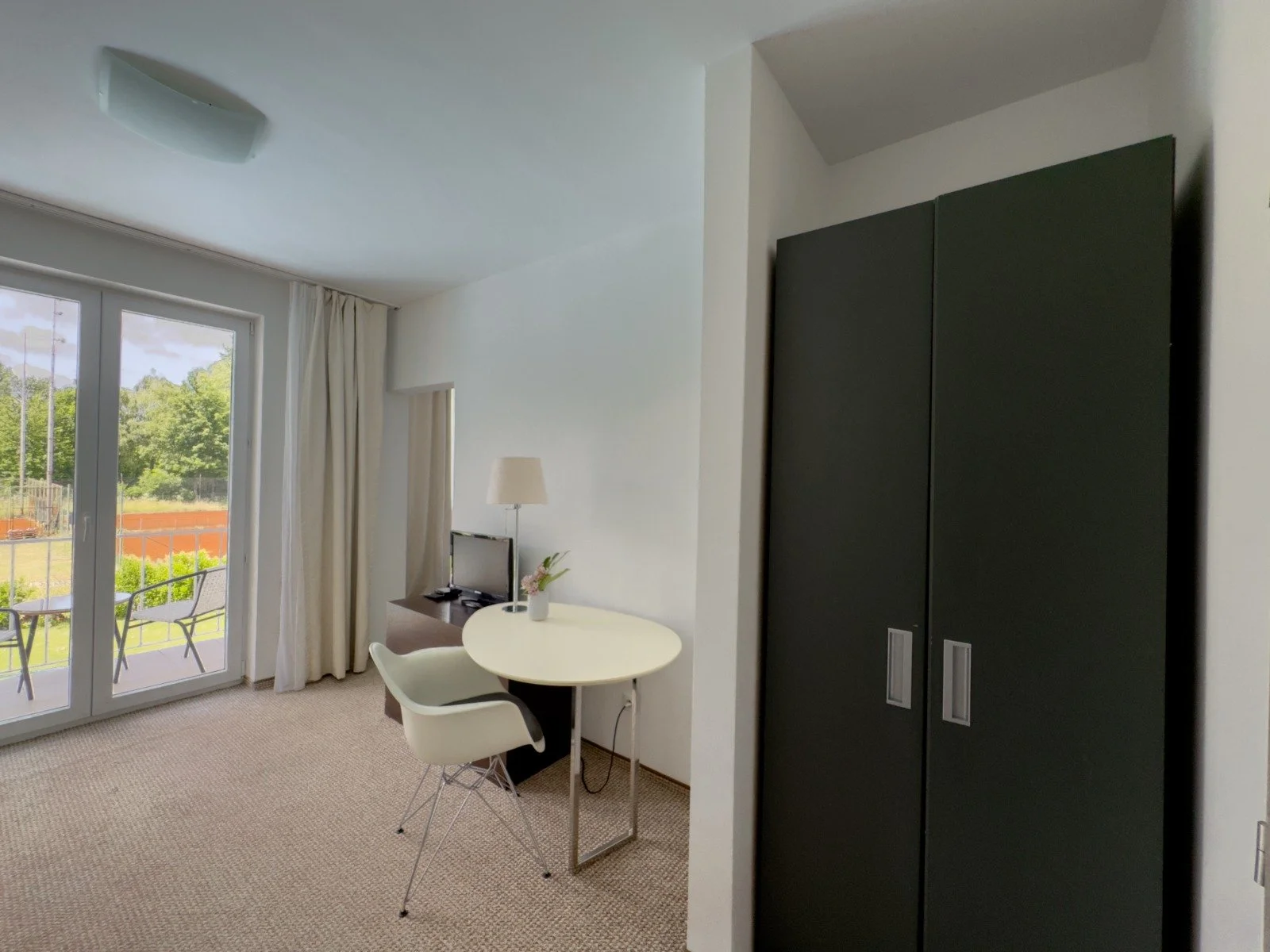 A living room with glass doors leading to a balcony, a small round table with a flower vase, a white chair, and a black wardrobe.