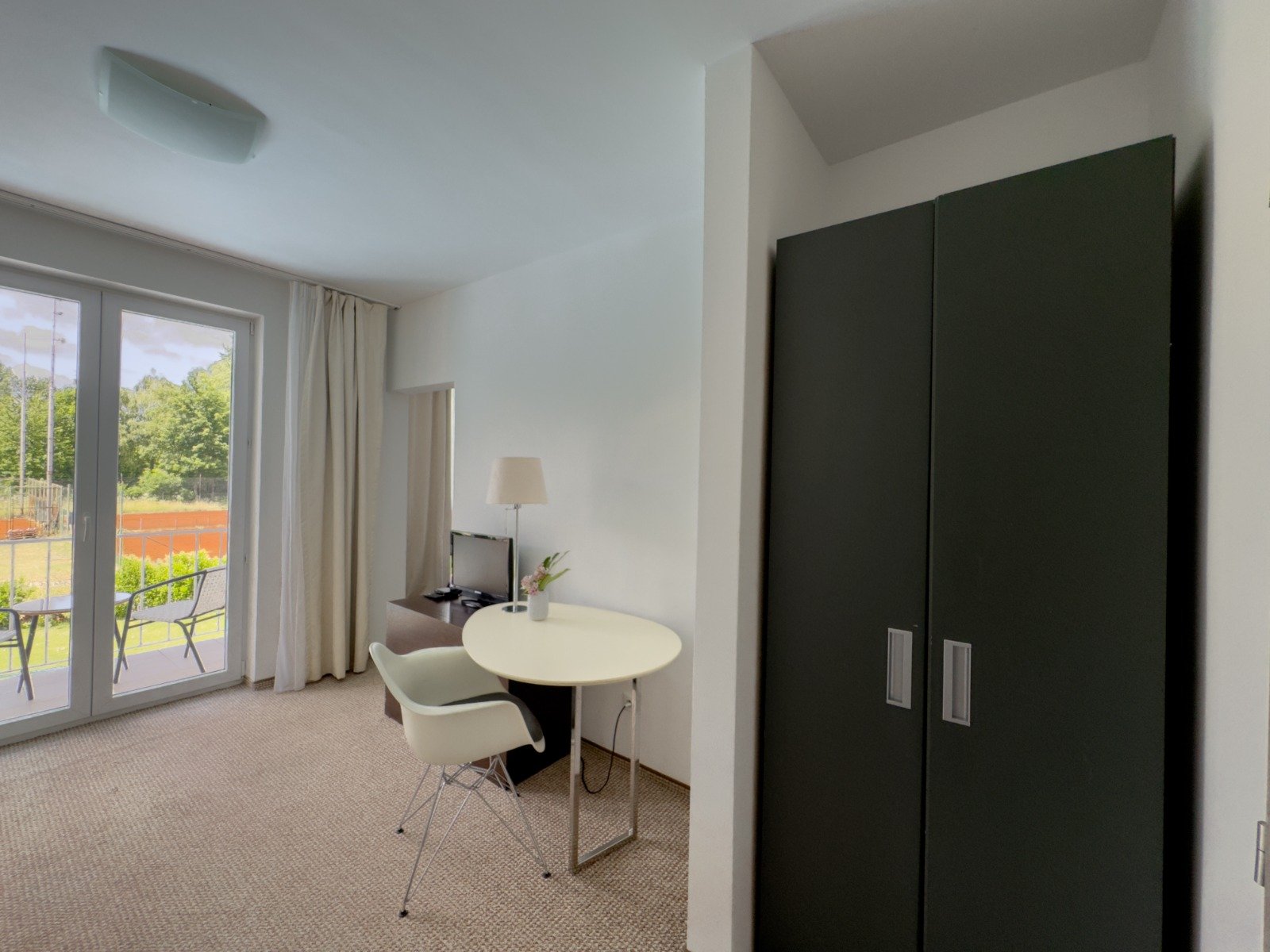 Living room with sliding glass doors leading to a balcony, small round dining table with a white chair, black wardrobe, TV on a dark wooden stand, and a lamp near the TV. Outside on the balcony are two chairs and a view of green trees.