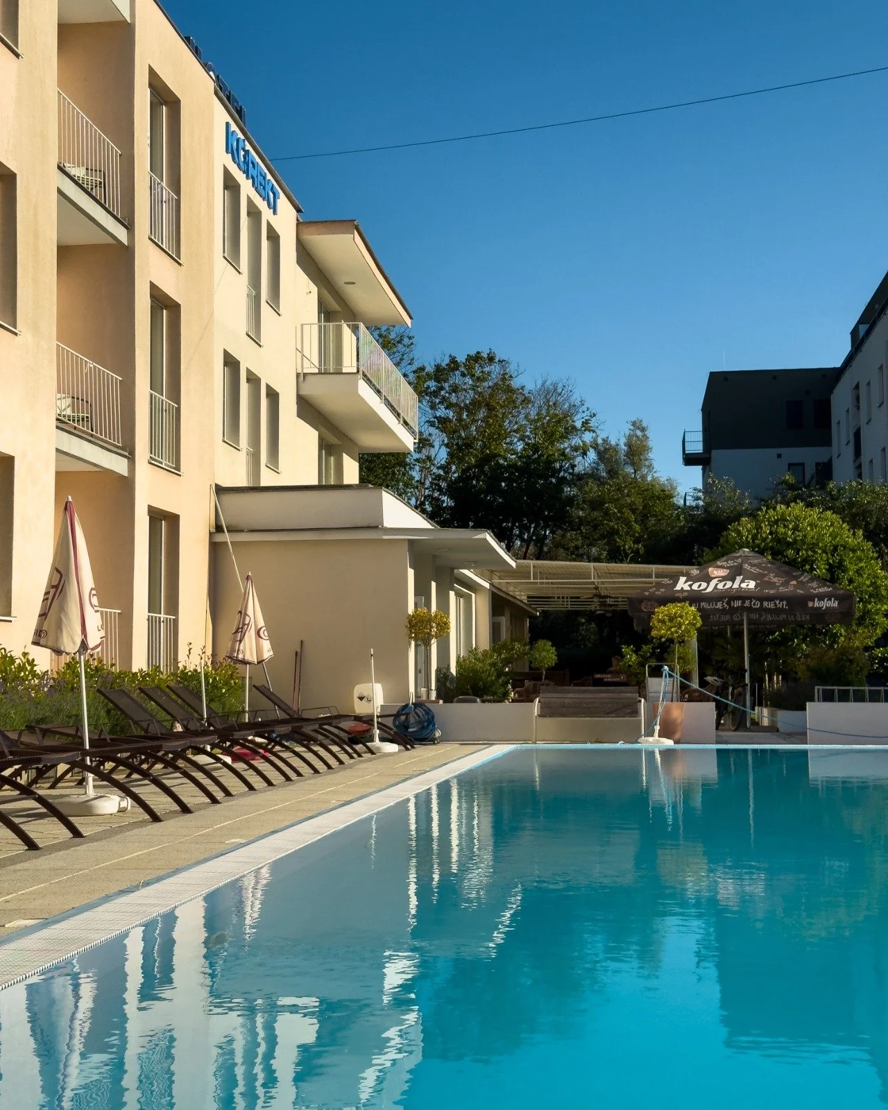 A hotel outdoor pool area with lounge chairs and umbrellas, modern apartment buildings in the background, under a clear blue sky.