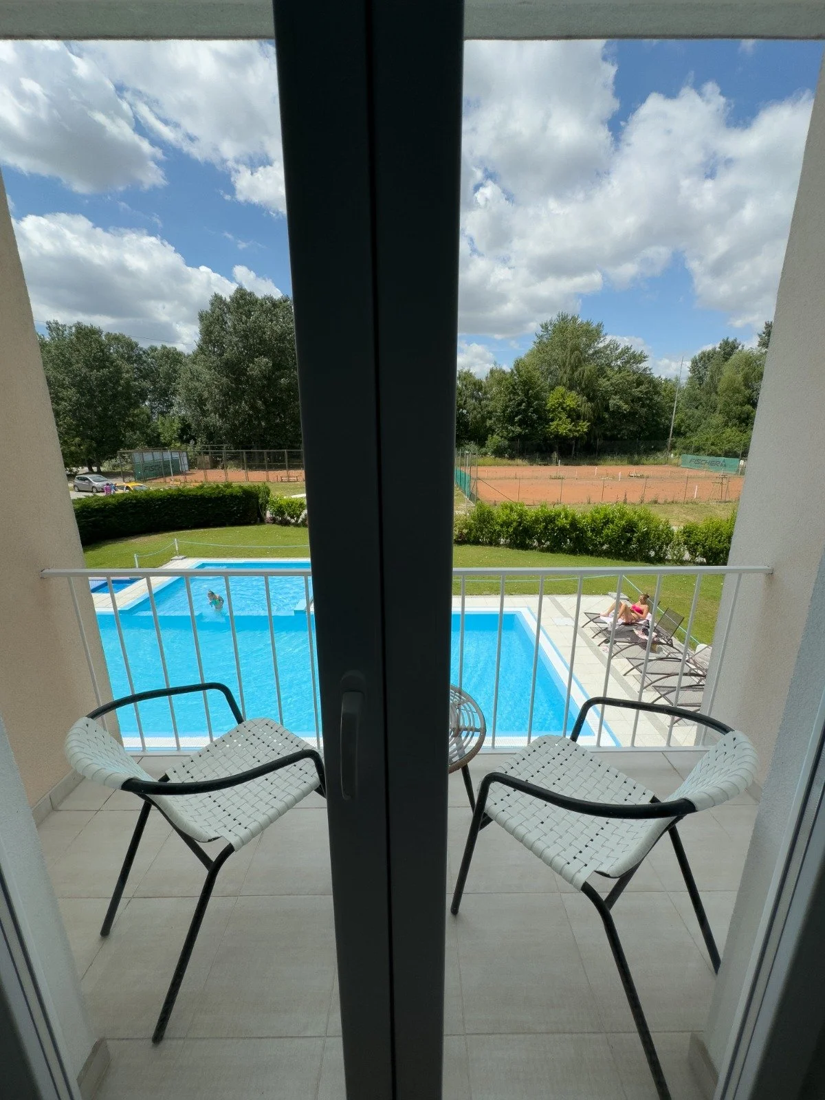 Balcony with two white chairs overlooking a swimming pool, a person swimming in the pool, and people lounging on chairs outside on a sunny day with cloudy sky.