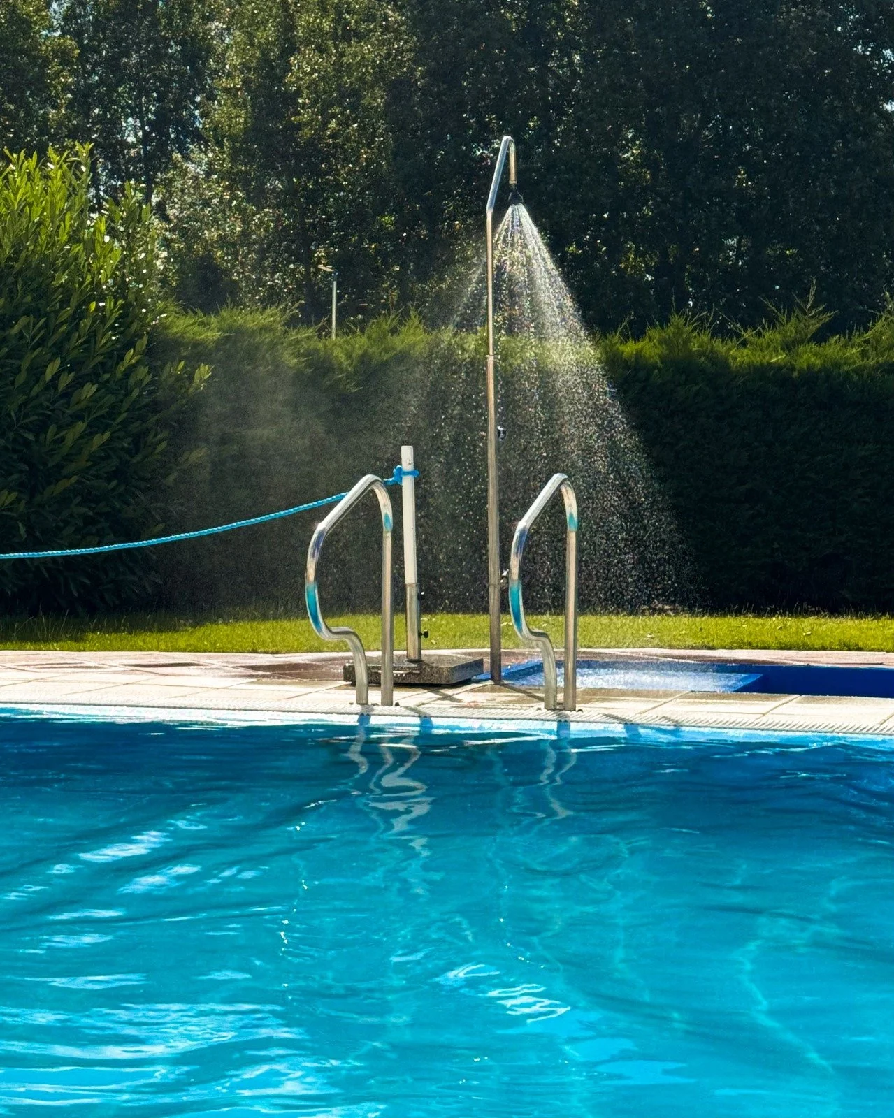 Public swimming pool with water fountain in the background, surrounded by green bushes and trees.