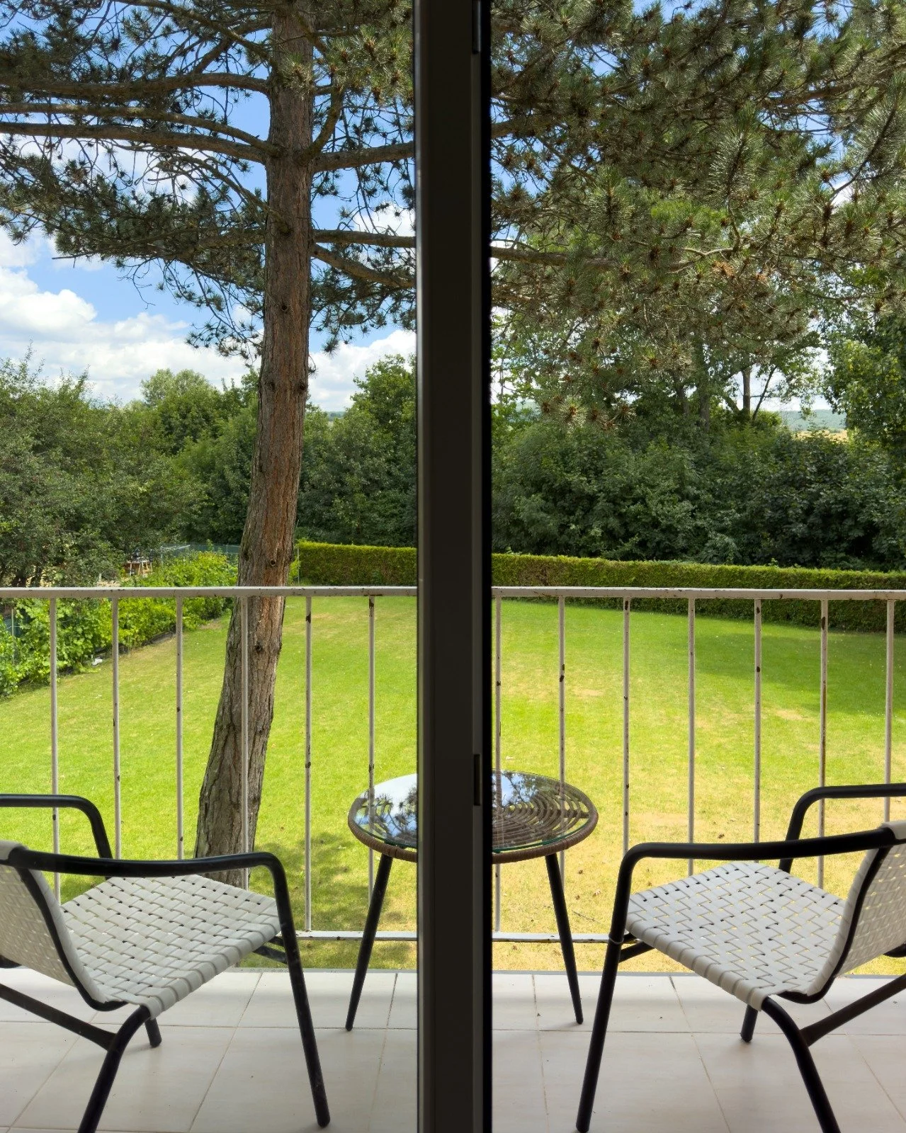 View through a sliding glass door showing a backyard with on patio, two white chairs, a small round table, a green lawn, trees, and a blue sky with clouds.