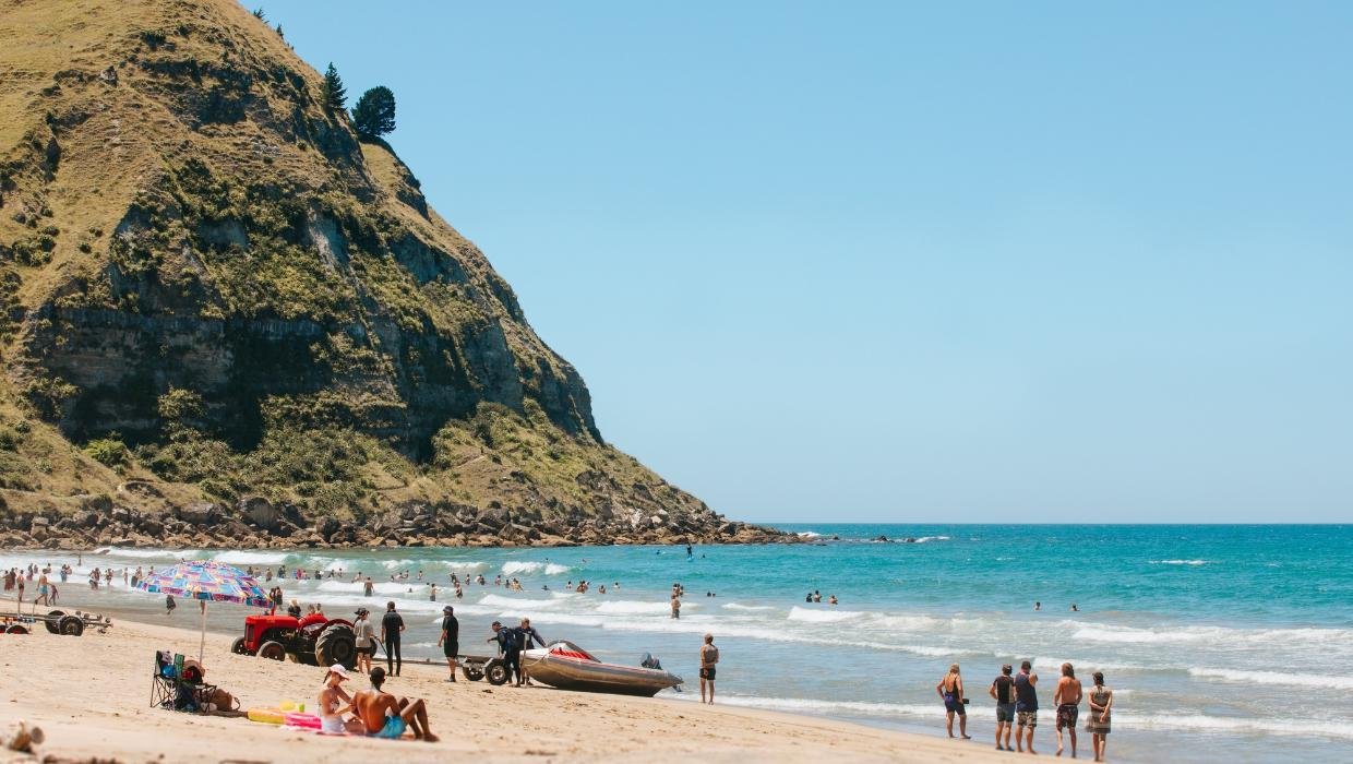 People enjoying a sunny day on a sandy beach with a large green hill in the background and turquoise ocean waves