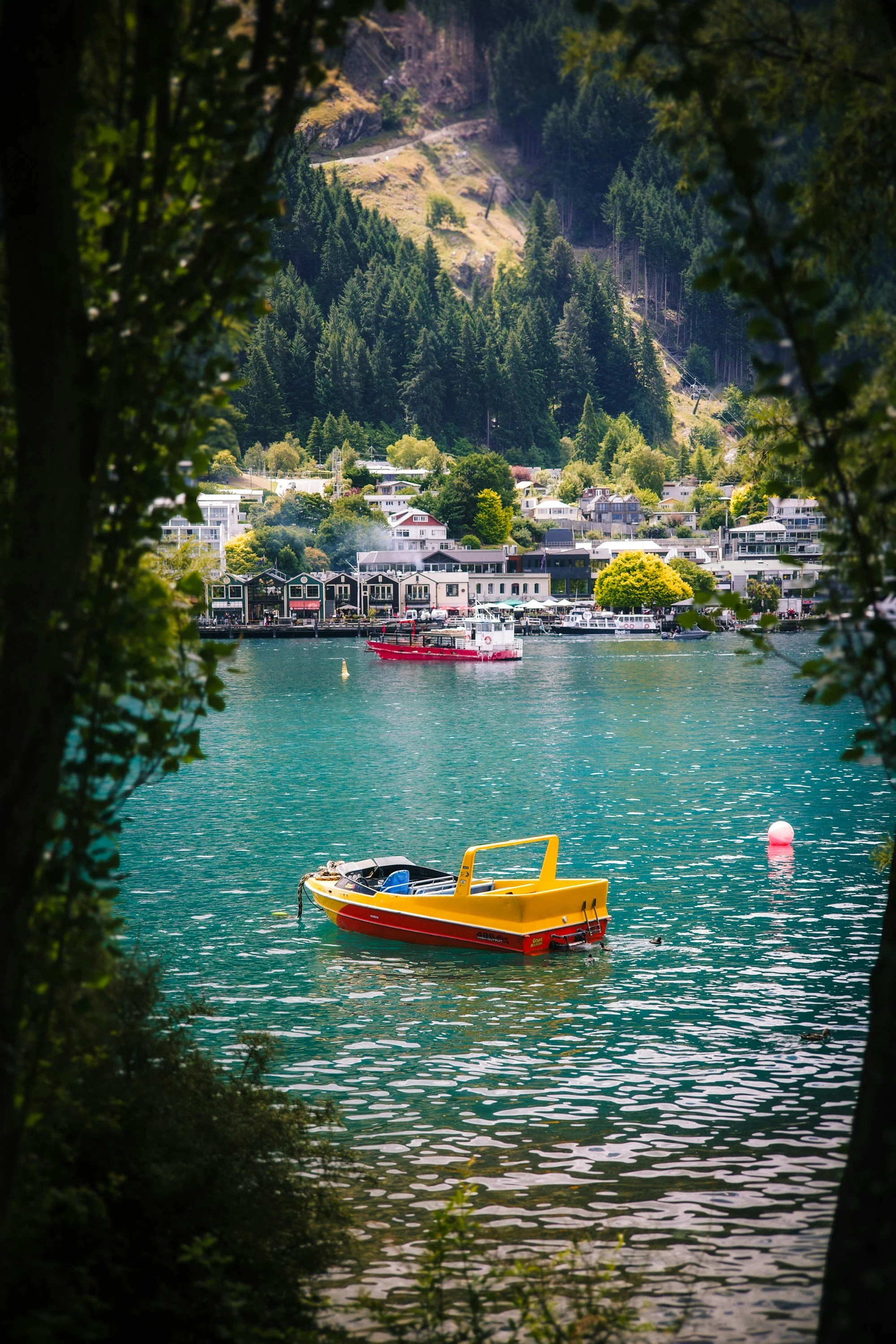 A scenic view of a small waterfront town nestled between lush green trees and a hill. A yellow and red boat floats on calm water in the foreground, with additional boats along the shoreline. The town features various buildings, including houses and commercial structures.