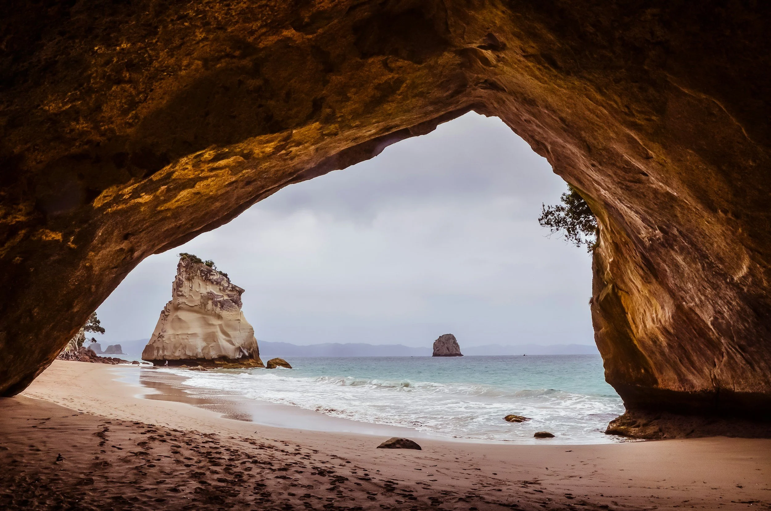 View through a large rocky arch on a beach, showing white cliffs and sea stacks in the distance.