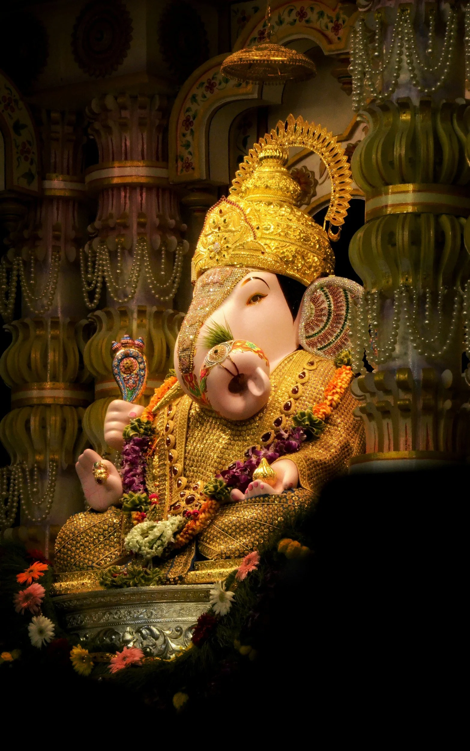 A decorative statue of Lord Ganesha, a Hindu deity, adorned with gold jewelry and a crown, surrounded by flowers and jewelry in a dark setting.