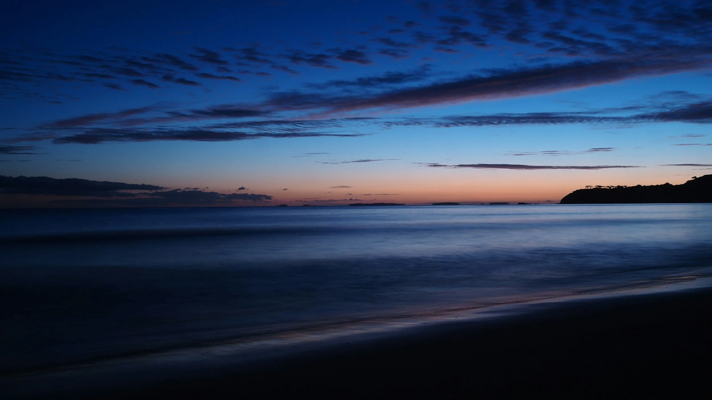 Calm ocean waters at dusk with a colorful sky and a silhouette of land in the distance.