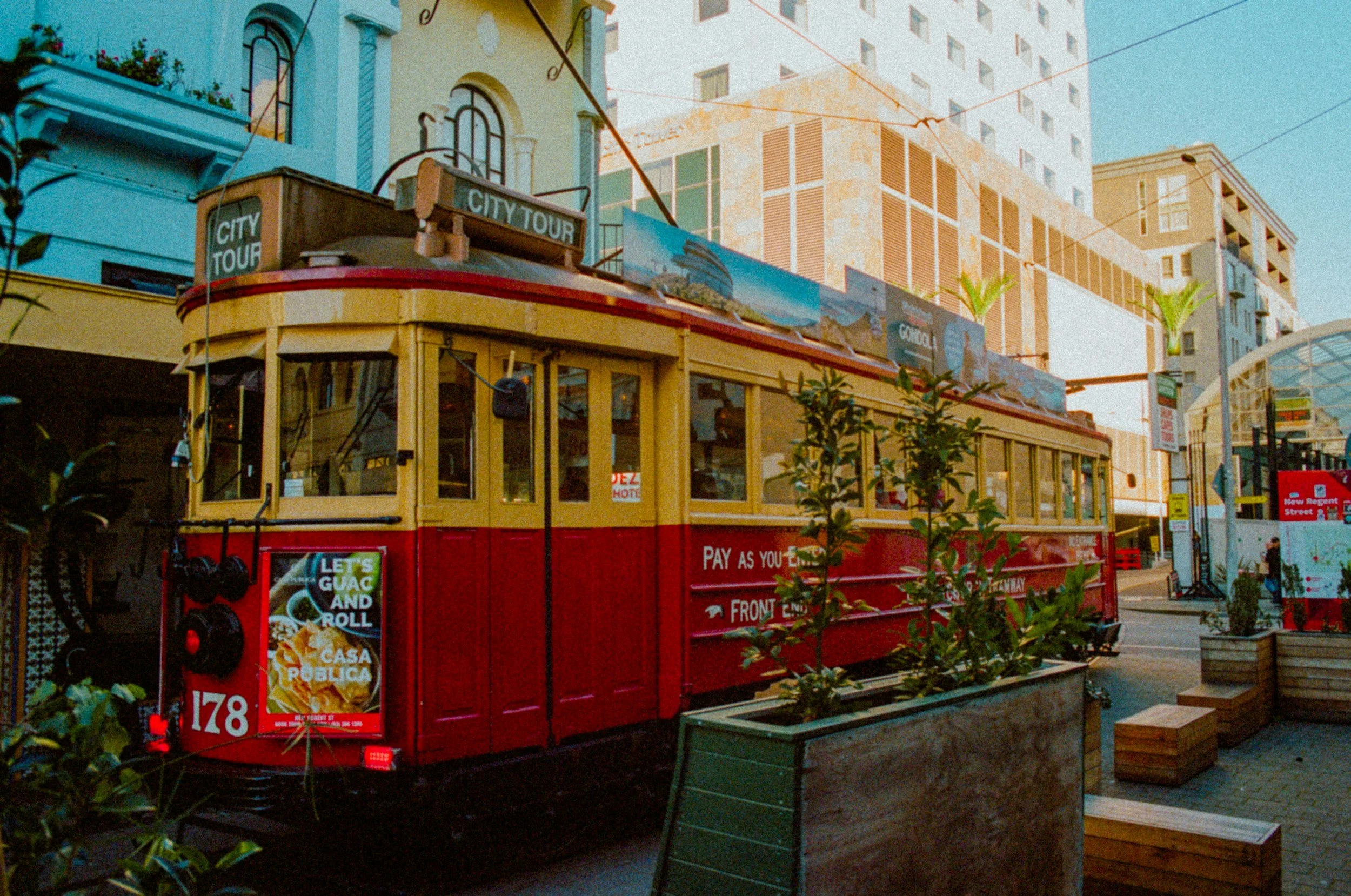 A red and yellow trolley with a sign that reads 'City Tour' on its roof. The trolley is parked on a city street with tall buildings and palm trees in the background. There is a sidewalk and some planters with greenery in the foreground.