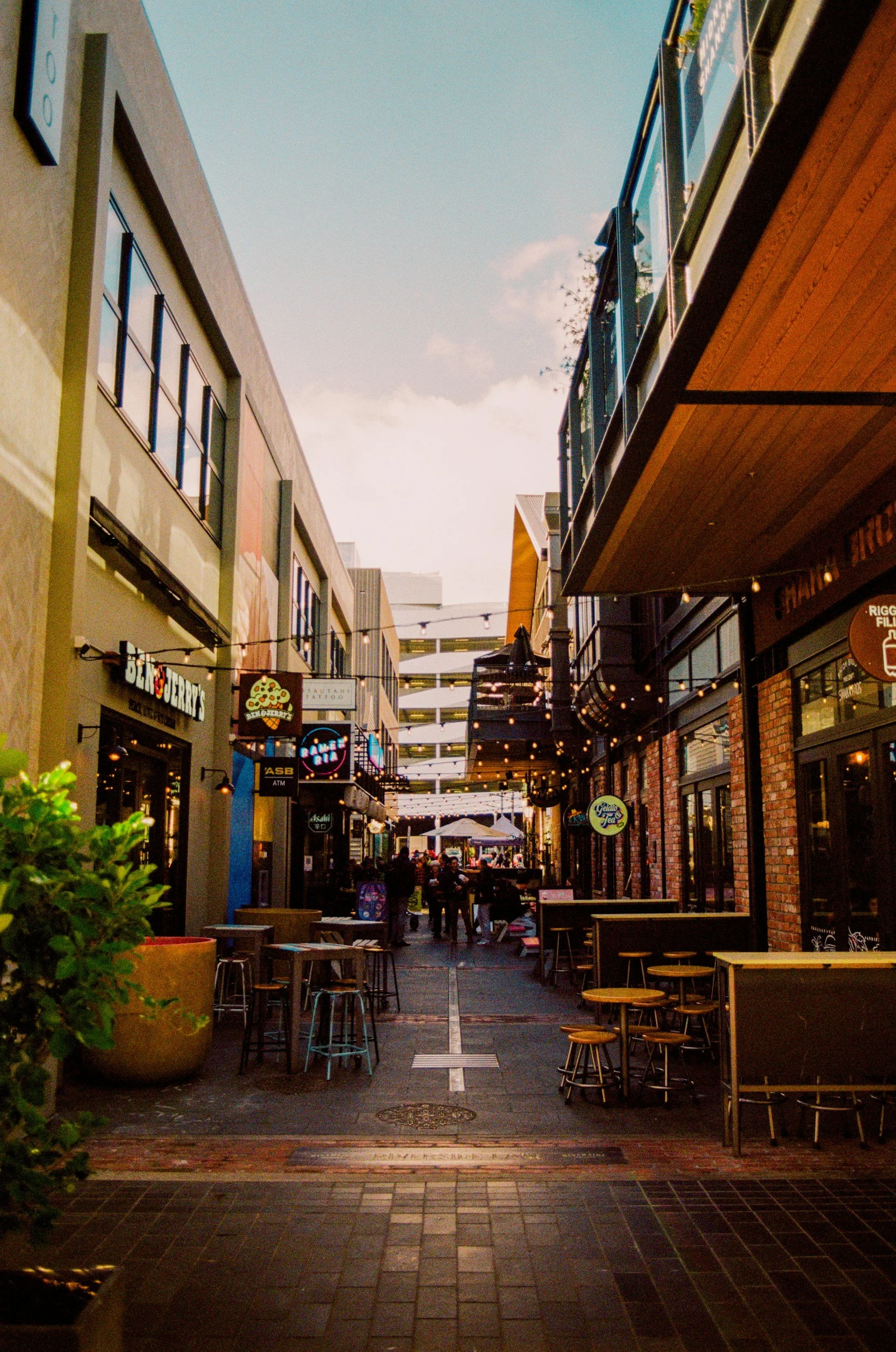 An outdoor shopping and dining area under a partly cloudy sky, with hanging string lights, various storefront signs, and people walking and sitting at tables.