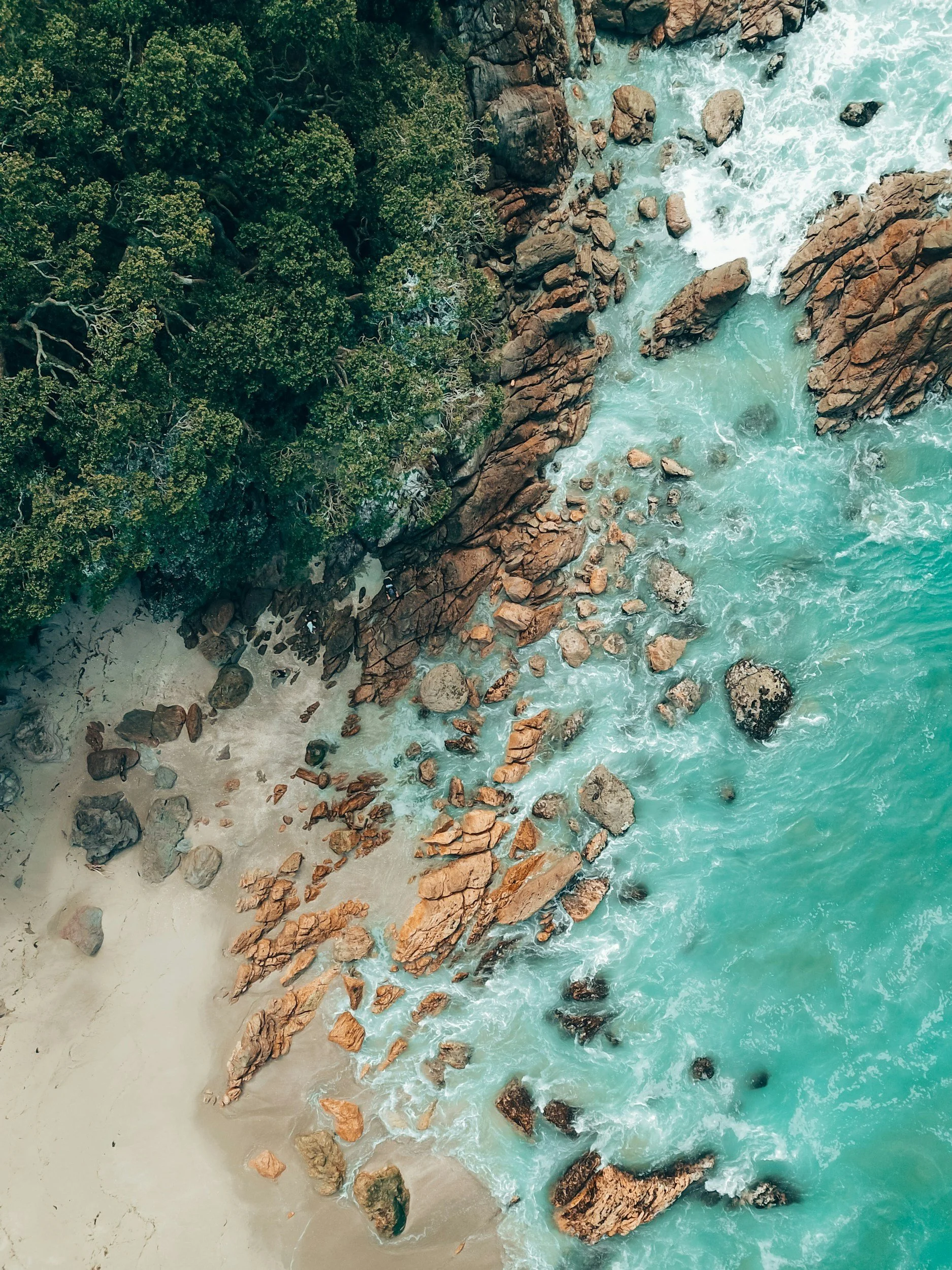 Aerial view of a coastline with green trees, red rocks, and turquoise ocean waves crashing against the rocks and sandy shore.