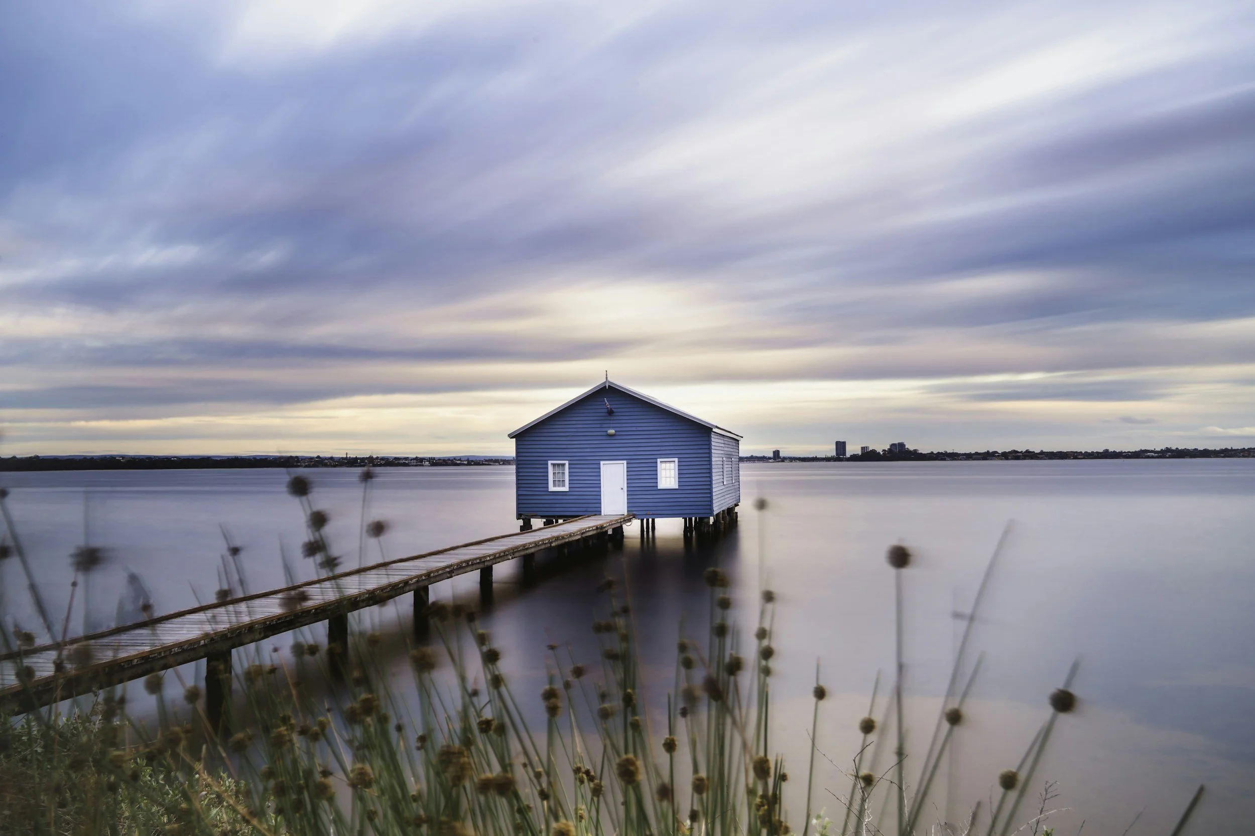 A small blue house on stilts over calm water, connected by a wooden walkway, with wildflowers in foreground and a cloudy sky overhead.