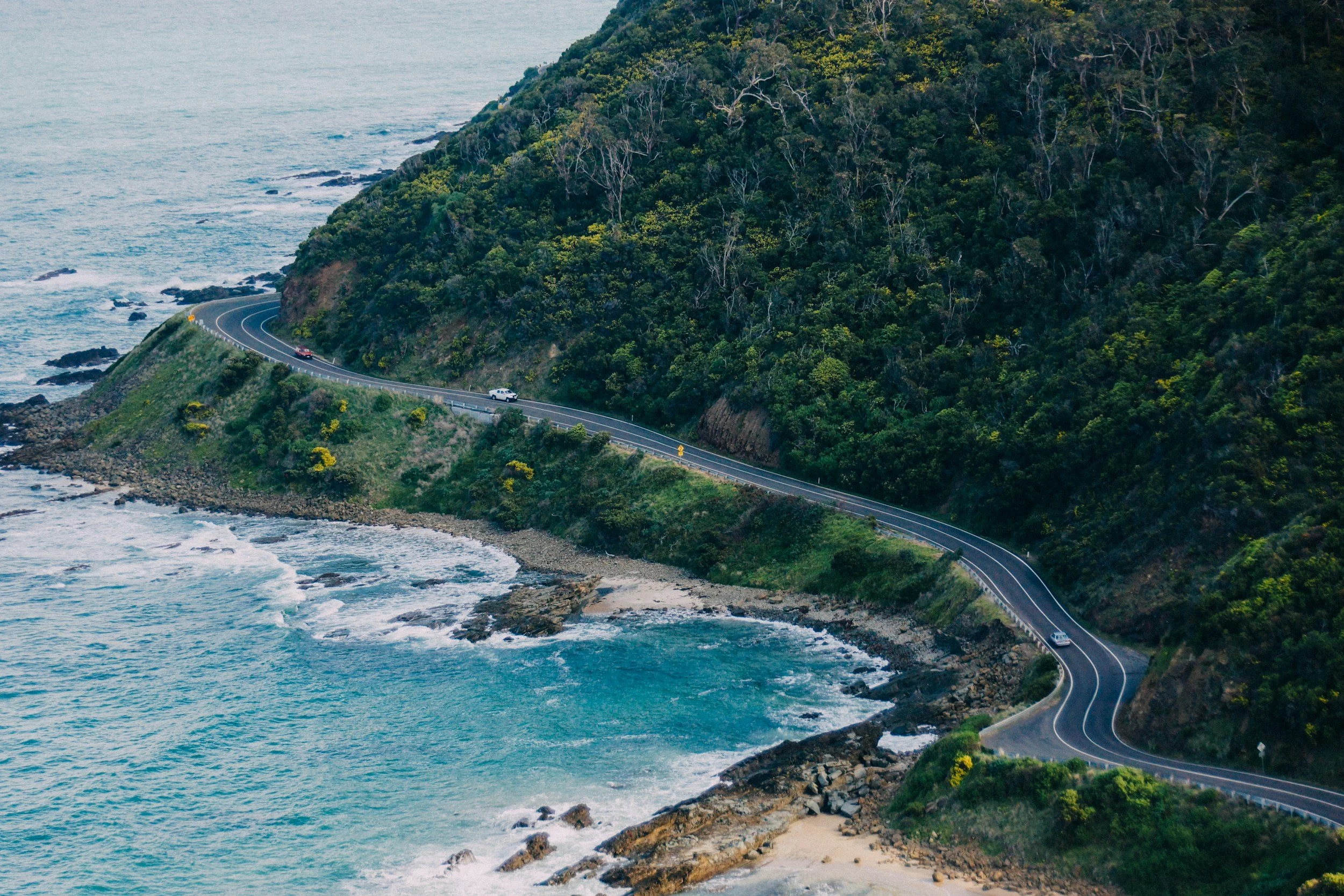 A coastal road winding along a hillside covered in green trees, with the ocean on one side and rocky shoreline below.
