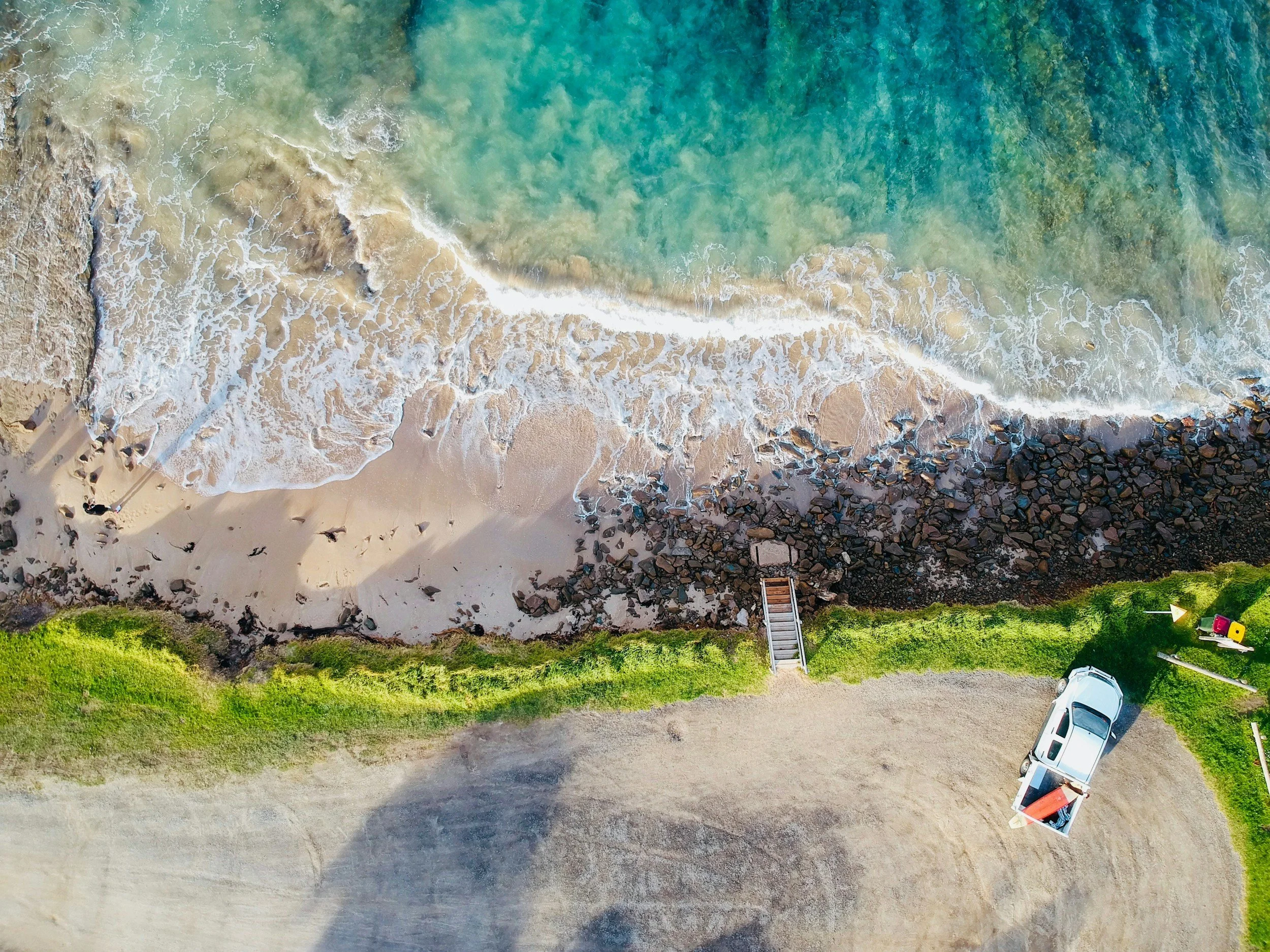 An aerial view of a beach with waves washing onto the sandy shore, rocky area on the right, green grass at the bottom, and a parking lot with several cars near the bottom right.