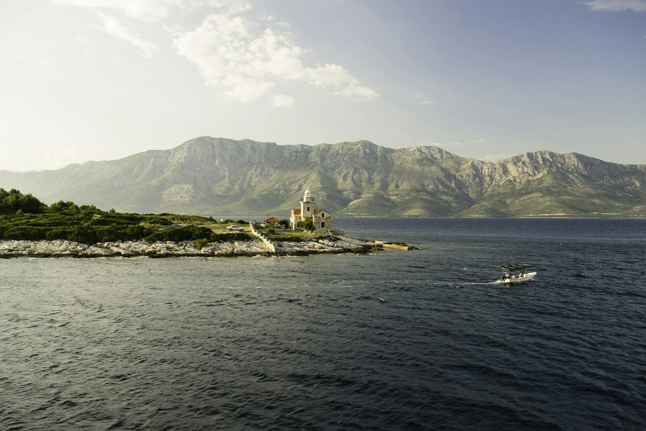 A scenic view of a coastal area with a lighthouse on a rocky outcrop, green shrubs, a boat on the water, and mountains in the background under a partly cloudy sky.